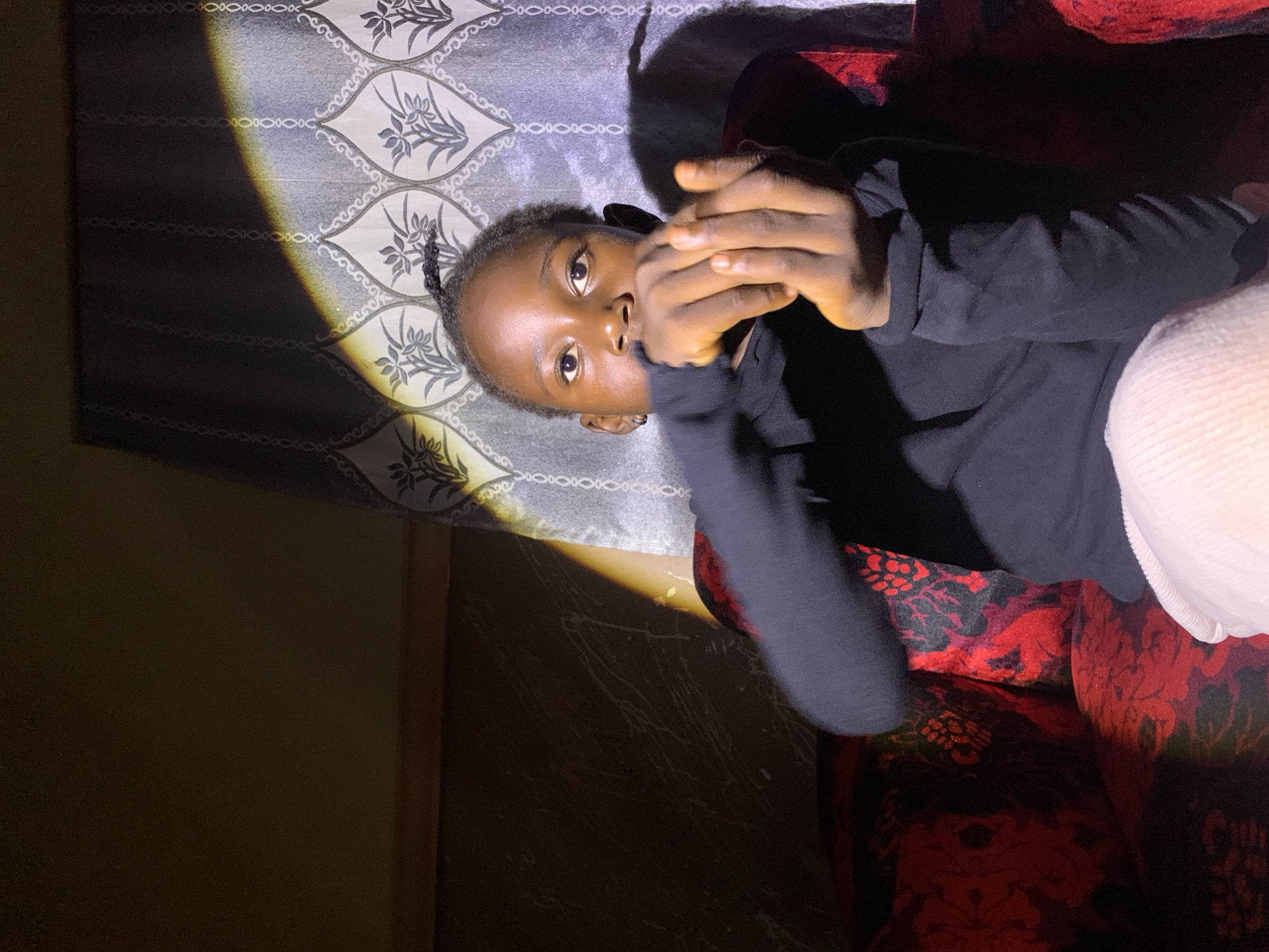 The image shows a young Black person sitting on a red and black patterned sofa. The person is wearing a black long-sleeved shirt and light-colored pants. They are holding their hands up in front of their face, with their fingers intertwined. A band of light from a window falls diagonally across the person and the patterned wallpaper behind them. The wallpaper has a repeating floral design in grey and white. The room appears to be dimly lit, with shadows present.