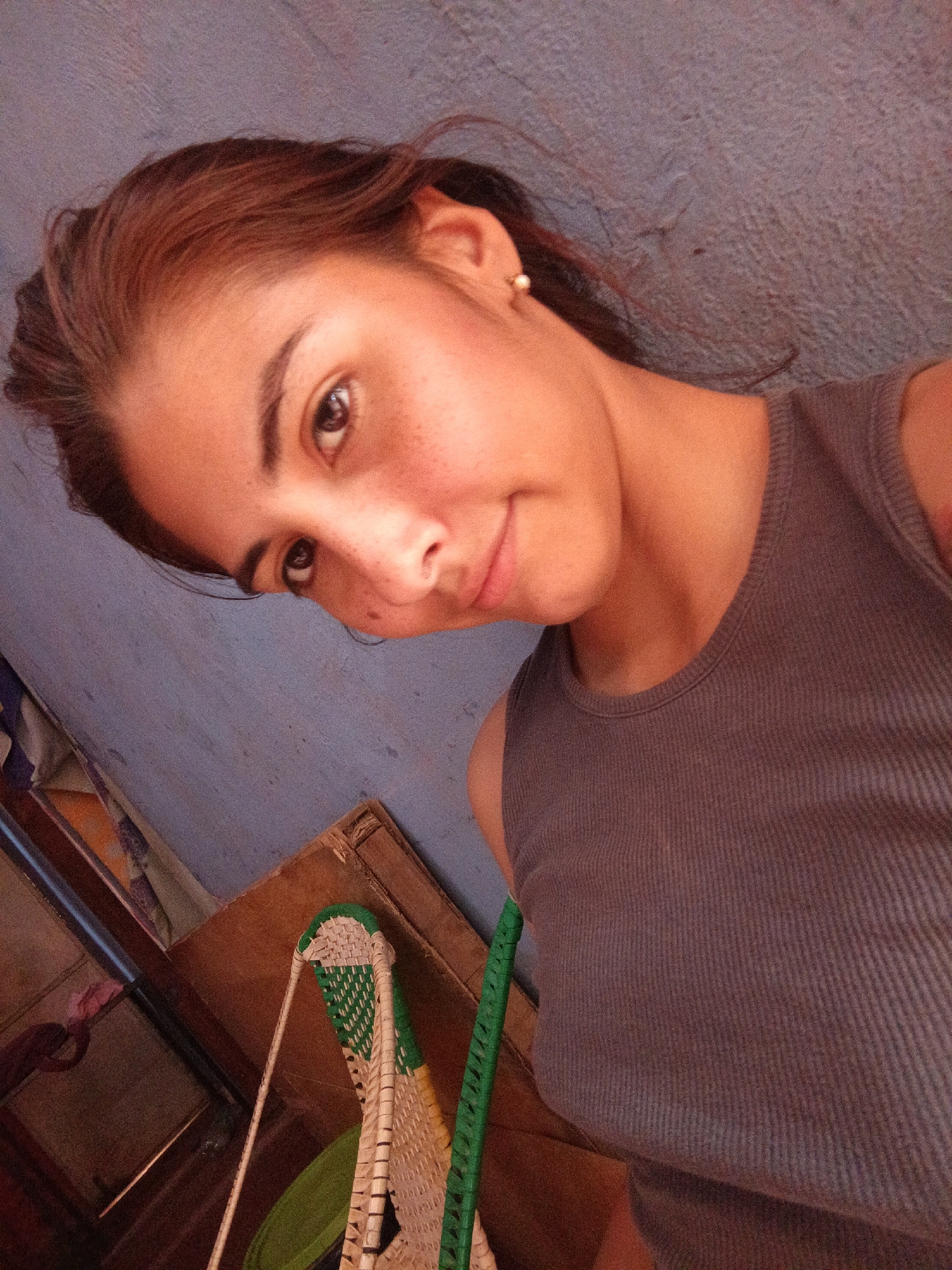 A young woman with light brown hair and brown eyes is shown from the chest up, angled downwards toward the camera, with a slight smile. She wears a gray ribbed tank top and a small earring in her left ear. She has freckles on her face. Behind her is a textured blue wall and parts of a wooden structure with green and white woven chair parts visible. Location: Unknown, Unknown.