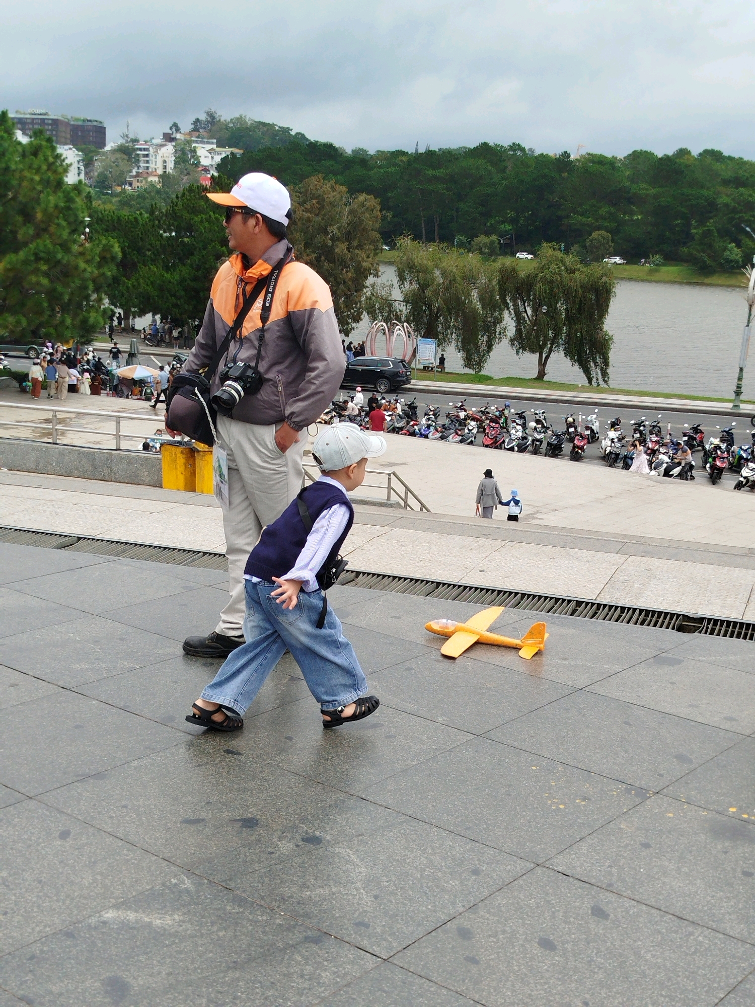 In Da Lat, Vietnam, a man in a white cap and orange-and-grey jacket stands to the left, holding a camera and bag. A young boy in a blue vest and jeans walks in front of him, looking to the right. A yellow toy airplane lies on the tiled ground in the foreground. In the background, a body of water is visible, with trees and buildings lining its shore. Several motorcycles are parked on a sidewalk further back.