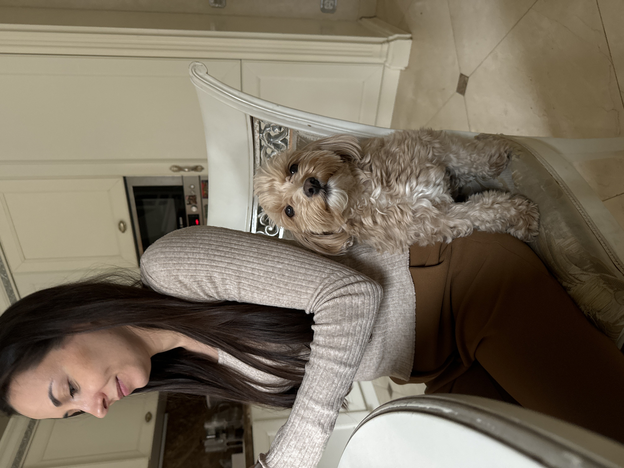 The image captures a woman with long, dark hair sitting in a chair. The woman is wearing a beige long-sleeved shirt and brown pants. A small, fluffy dog with light brown fur rests on the back of the chair behind her. The setting appears to be inside a kitchen, with light-colored cabinets and an oven visible in the background. The specific location is Krasnoyarsk, Russia.