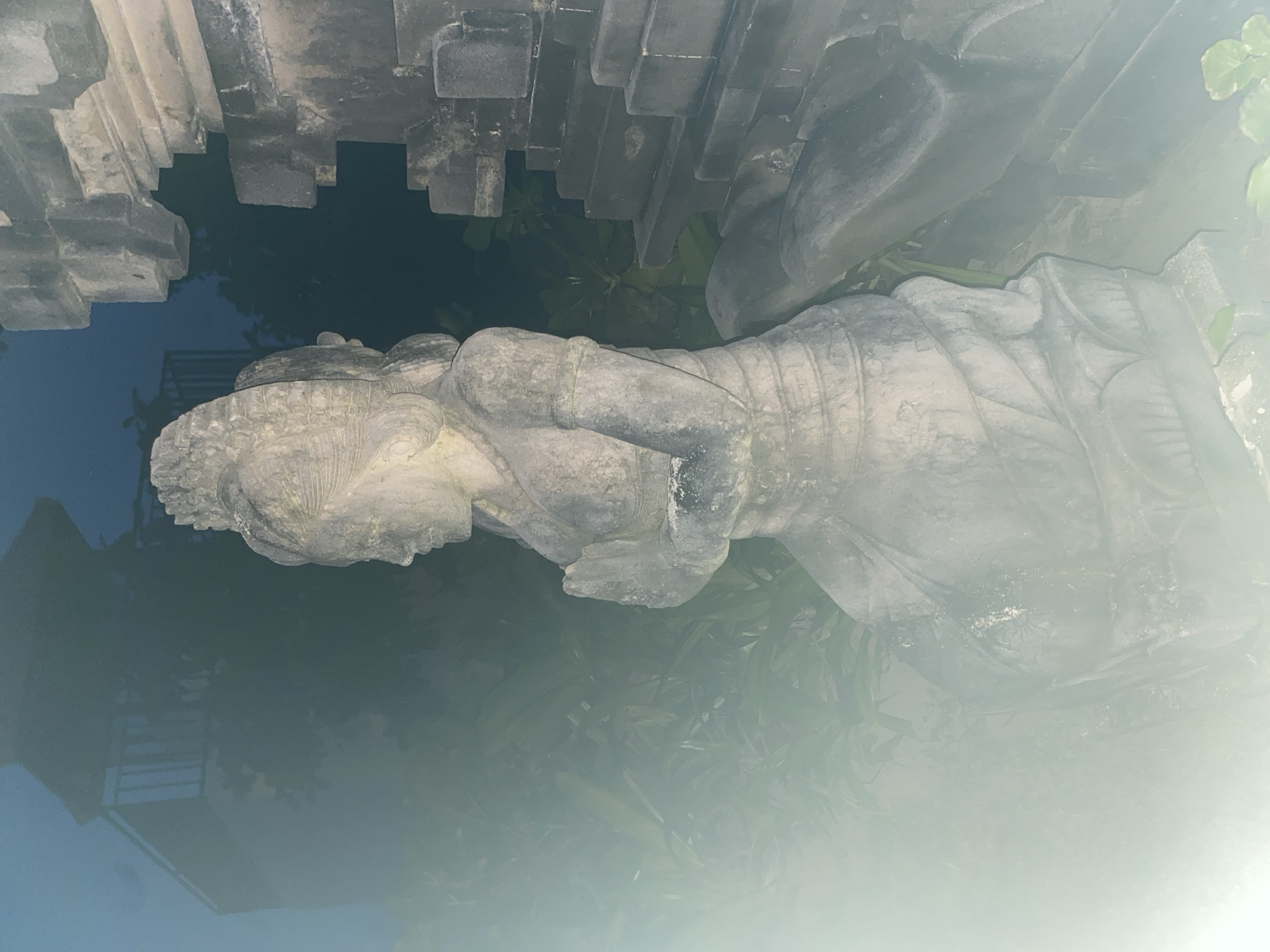A horizontal photograph depicts a stone statue next to a tiered stone structure with foliage in the background. The image is oriented upside down.
The statue is weathered and appears to be of a person lying down, possibly sleeping. The figure wears some type of head covering and is dressed in what appears to be a garment wrapped around the torso. The stone structure has a tiered design and appears to be part of a building or monument. Foliage is visible behind the statue and structure. The background is slightly obscured by haze or mist.
Based on the metadata and additional information, the image was taken in the city of Kuta Selatan, Indonesia.