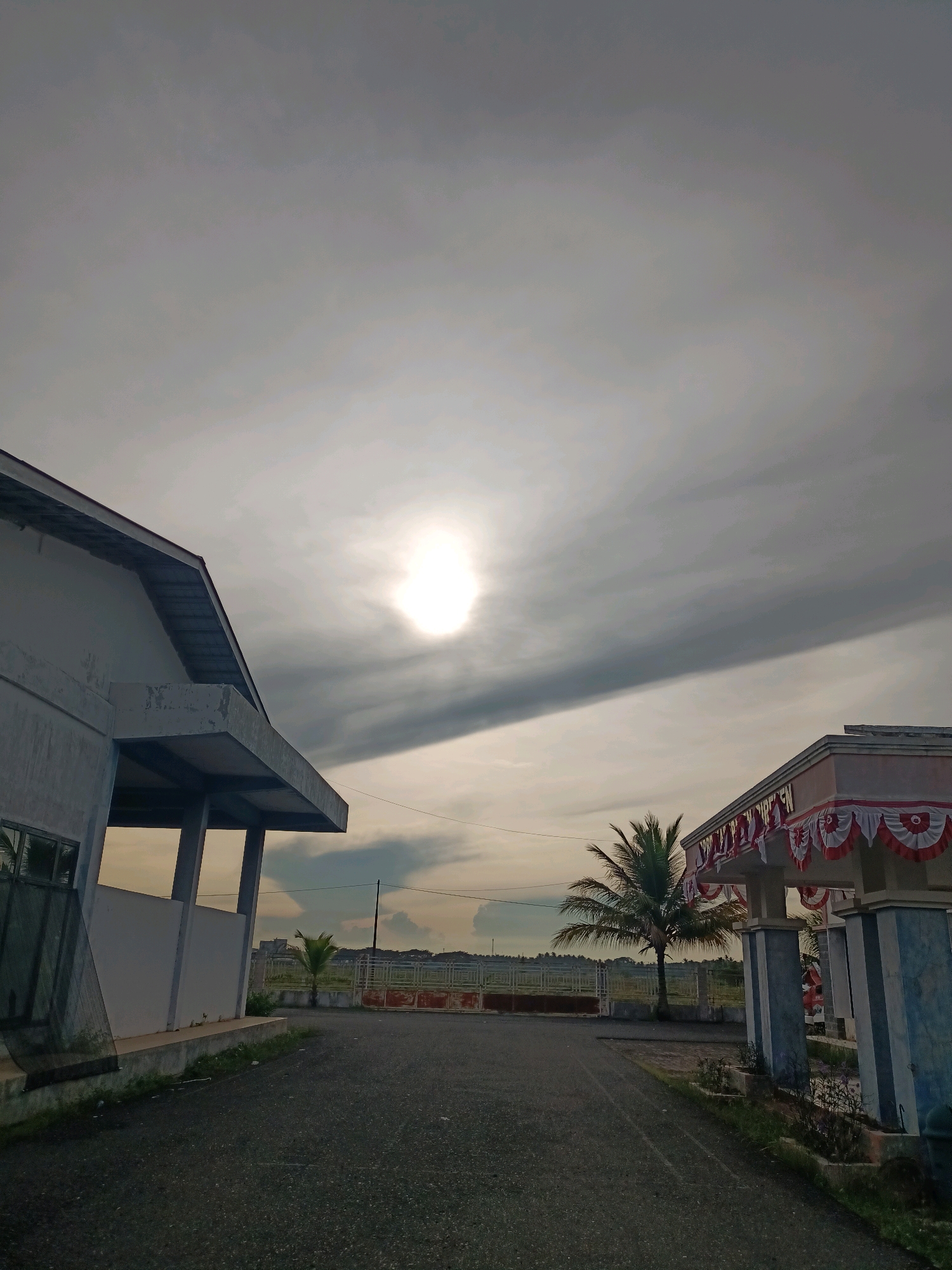 The image depicts a view of a building and its surroundings in Blangbladeh, Indonesia, under an overcast sky with the sun visible as a bright orb. On the left, a white building with a gray roof and overhang is partially visible, extending from the left edge of the frame towards the center. To the right of the frame, another structure with blue columns and a white roof adorned with red and white decorative bunting is present. A paved asphalt path leads from the foreground into the distance between these two buildings. A palm tree stands between the buildings, with a fence and a field beyond. The sky is filled with heavy, dark gray clouds, creating a diffused light across the scene. The overall impression is a quiet, possibly early morning or late afternoon scene.