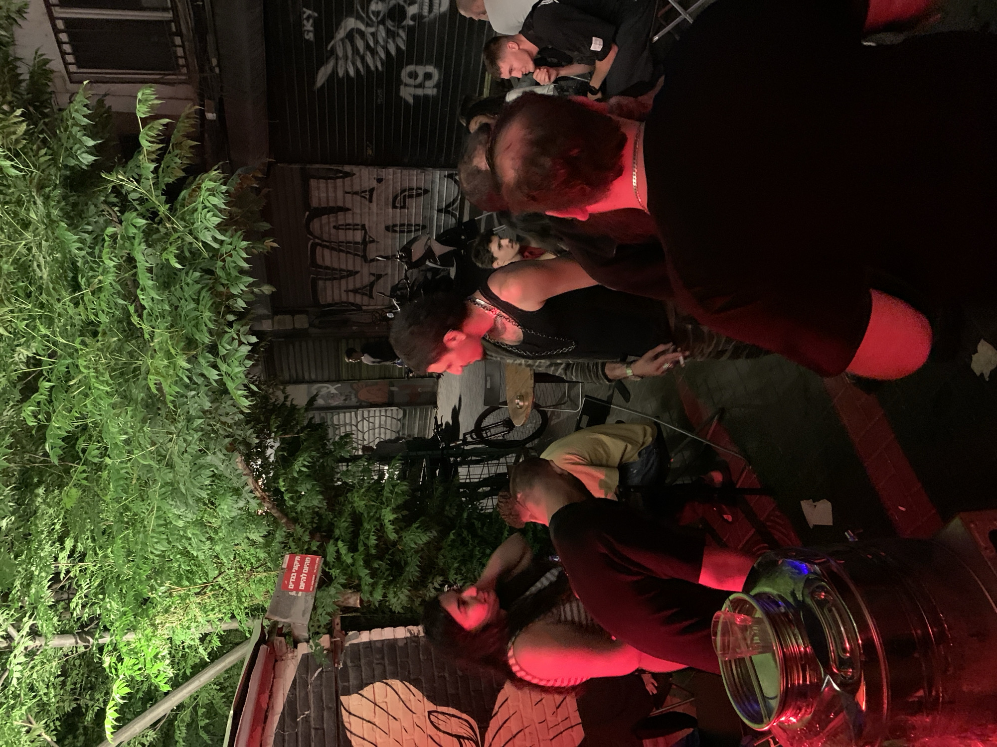 The image portrays a lively night-time outdoor gathering, likely a social event or a designated area of a bar or establishment in Tel-Aviv, Israel. The scene is illuminated by strong artificial lighting, predominantly red and green, creating a distinct ambiance against the overall darkness.
In the immediate foreground, several individuals are visible. A person with dark hair, wearing a black tank top and a silver chain necklace, is prominently featured, their face and upper body illuminated with a vivid red glow. Another person, seen from the back in a dark shirt, is also bathed in red light. To the far right, a large, reflective beverage dispenser, possibly glass or polished metal, catches both red and blue light, suggesting it contains drinks.
The background shows more people socializing, standing and conversing, contributing to a casual and vibrant atmosphere. Green foliage, resembling dense bushes or trees, occupies a significant portion of the left side of the frame, brightly lit by green lights. Behind the gathering, buildings or storefronts with closed metal shutters are visible. These shutters feature graffiti, including the word "SKY" and the number "19", along with other less legible markings. Bicycles are also faintly discernible, parked in the background.
A notable detail is a red sign mounted on a pole amidst the greenery on the left. The sign displays Hebrew text, which translates to: "No entry with outside drinks and glass containers." This specific instruction indicates the location is a commercial venue with rules regarding external beverages. The combination of artificial lighting, graffiti, and the casual gathering points to a popular nightspot.