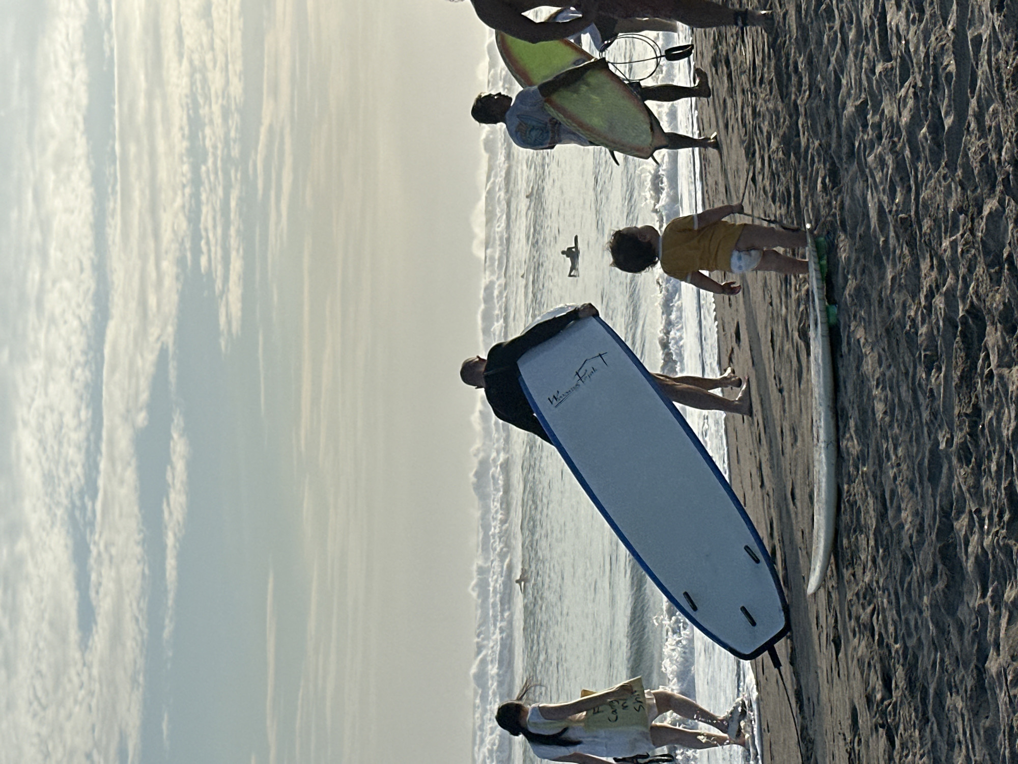 A group of people are on a sandy beach in North Kuta, Indonesia, with surfboards. In the foreground, a young child wearing a diaper stands facing away from the camera, holding a leash attached to a surfboard. To the left, a woman in a white dress walks towards the water, carrying a bag with writing on it. A large blue and white surfboard is being carried horizontally by a person whose torso is visible. To the right, another person is carrying a yellow and green surfboard, and further to the right, a person's legs are visible. In the background, waves are crashing, and several surfers are in the water. The sky is overcast with wispy clouds.