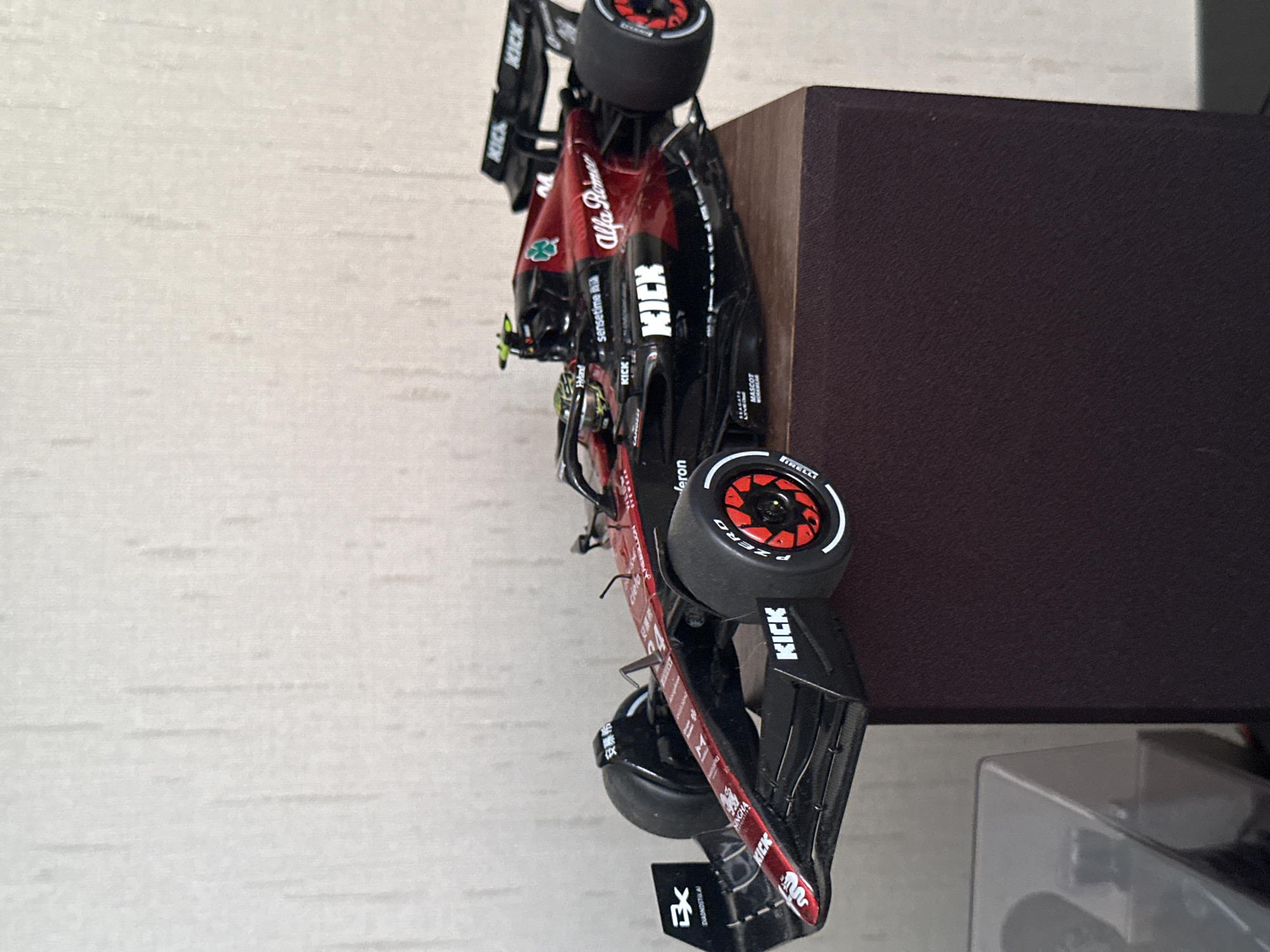A detailed, forensic, matter-of-fact description of the scene is not possible as the image does not contain information that would allow for the identification of the city of Krasnodar, Russia. The image shows a model of an Alfa Romeo Formula 1 race car, predominantly black and red with various sponsor logos. The car is positioned on top of a dark brown, rectangular object, possibly a speaker or a piece of furniture. The background is a textured, light-colored wall.