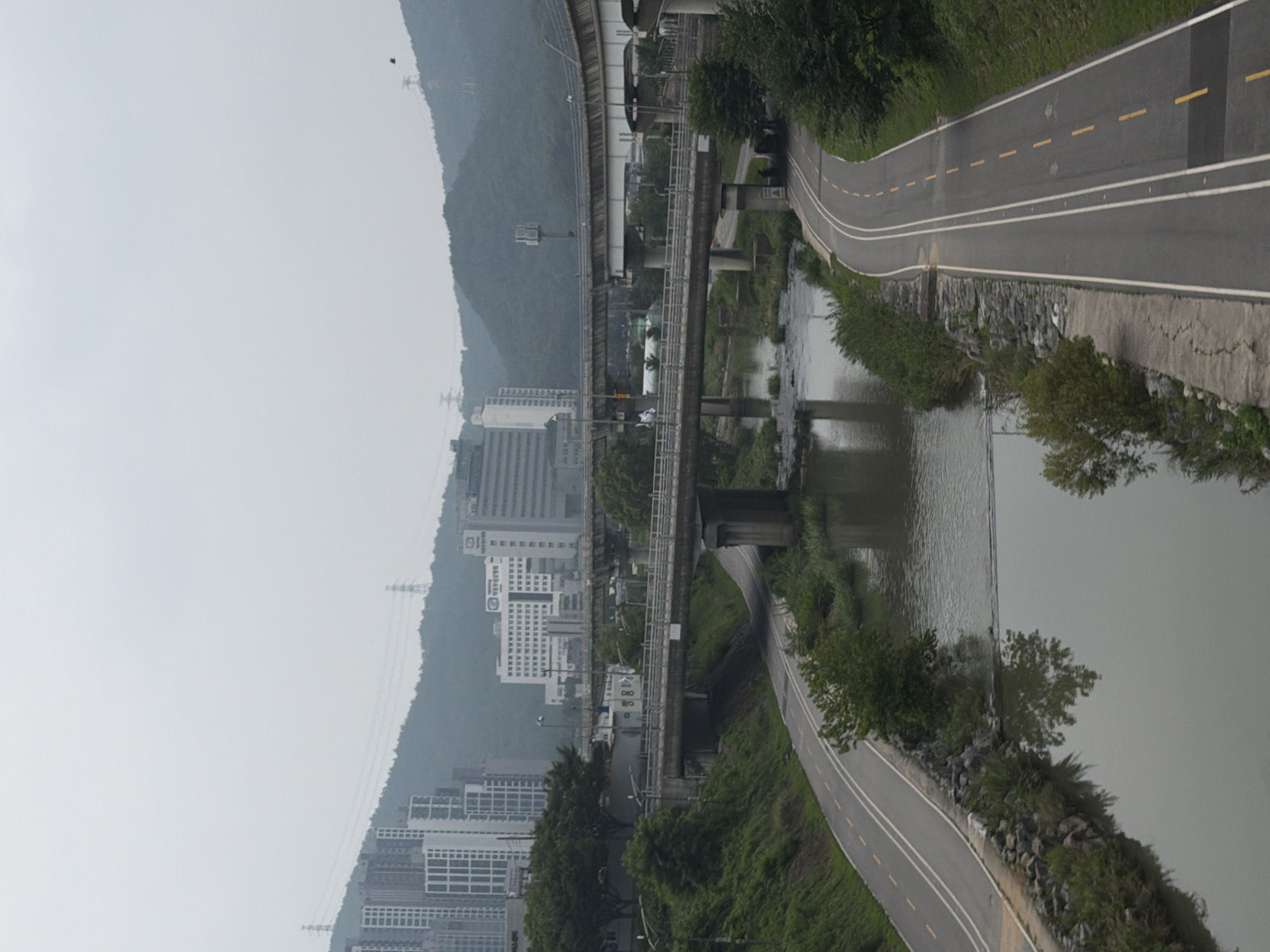 This media file, oriented vertically, showcases a somewhat overcast day in Uijeongbu-si, South Korea. The scene features a curving asphalt road with white and yellow lane markings, bordering a body of water that reflects the muted sky. Lush green vegetation lines the road and the water's edge, interspersed with sections of grey stone embankment.
In the background, a sprawling urban landscape unfolds against a misty, mountainous backdrop. Several tall buildings, primarily residential or office complexes, are visible, characterized by their modern architecture and numerous windows. Elevated infrastructure, likely an overpass or a train line, runs parallel to the road and water, supported by concrete pillars. The overall atmosphere is tranquil and somewhat muted due to the overcast weather. No people are clearly visible in the frame. Visible text includes a sign on one of the buildings that appears to read "MANSION" and another smaller sign on the elevated structure with Korean characters.