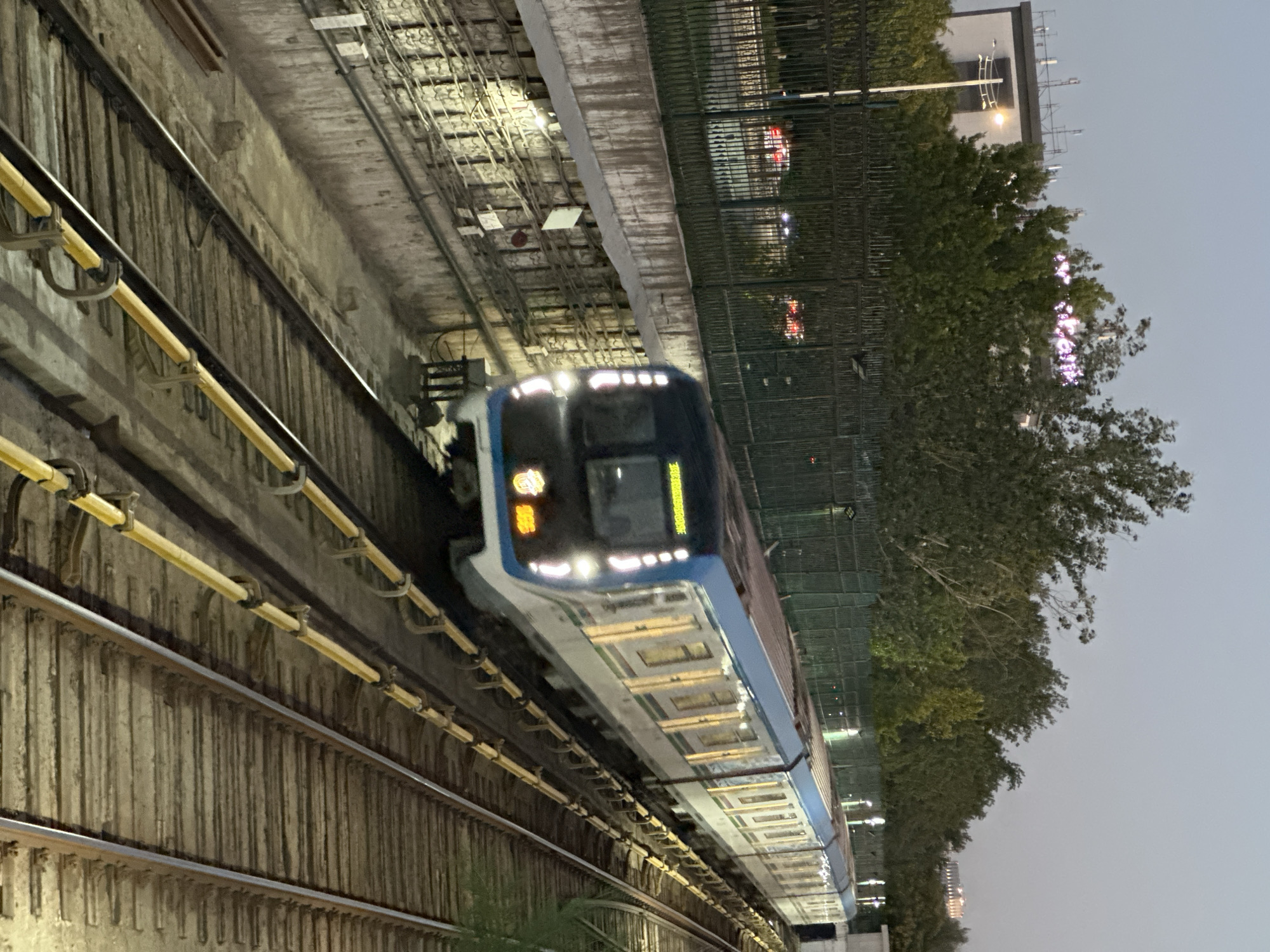 An elevated metro train with blue and white carriages is traveling on a track. The train has its headlights on and appears to be in motion, indicated by the blur. The train has windows lit from within, suggesting passengers are inside. The tracks are lined with yellow safety barriers. On the left, a concrete wall is visible with electrical conduits and lighting fixtures. Beyond the track, there's a fence and trees, and in the distance, buildings and a hazy sky are visible. The scene suggests an urban environment, possibly at dusk or dawn. The cropped image shows a building with illuminated text, which reads "Yandex", a company with operations in Uzbekistan. This suggests the location is likely Tashkent, Uzbekistan, given Yandex's presence and the architectural style.