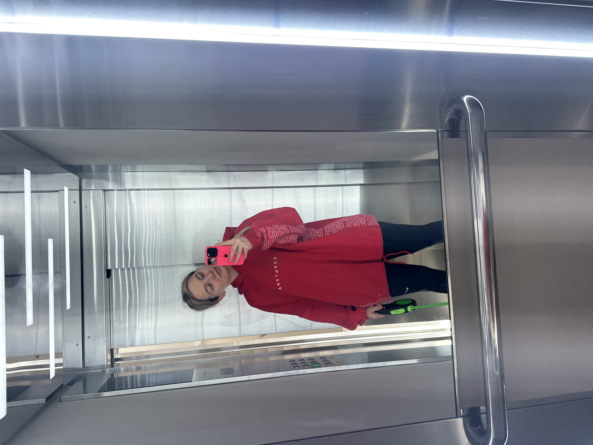 The image depicts a woman taking a mirror selfie in an elevator. She is wearing a red hooded sweatshirt with white lettering and black leggings. She is holding a pink smartphone. A retractable dog leash, with a green and black handle and green leash, is visible in her left hand. The elevator has brushed metal walls and ceiling with integrated lighting. Elevator control buttons are visible. The location is potentially in Budva, Montenegro, based on possible reflections or the general type of architectural elements.