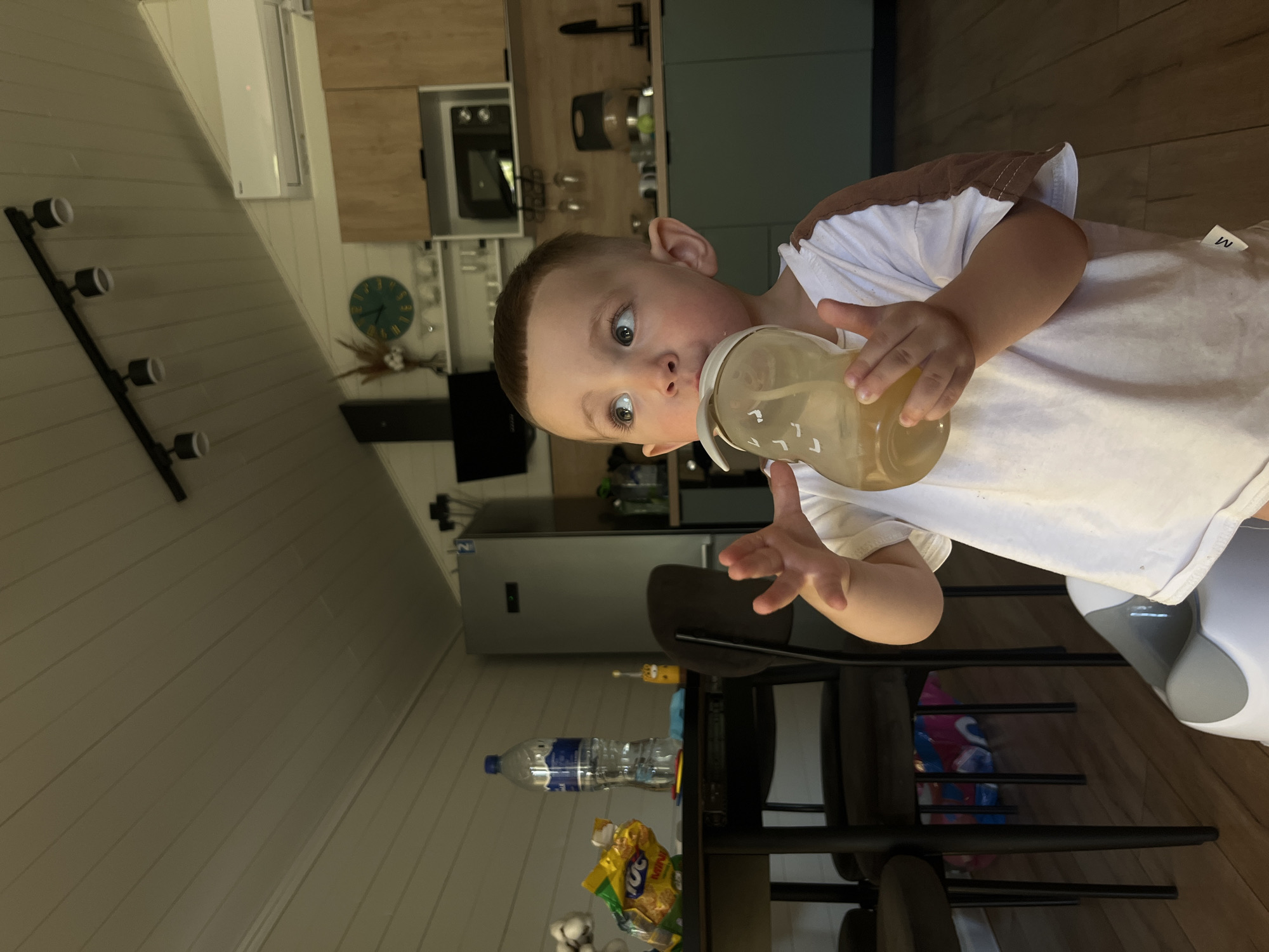 A young child with fair skin, light brown hair, and large eyes drinks from a clear bottle containing a light-yellow liquid. He wears a white T-shirt with a brown trim. The child is positioned in a kitchen setting. A dark metal chair is nearby. On a table are a bottle of water, a bag of snacks, and some cotton balls. A refrigerator, kitchen cabinets, and a microwave are visible in the background. The location is possibly Литвиново, Russia.