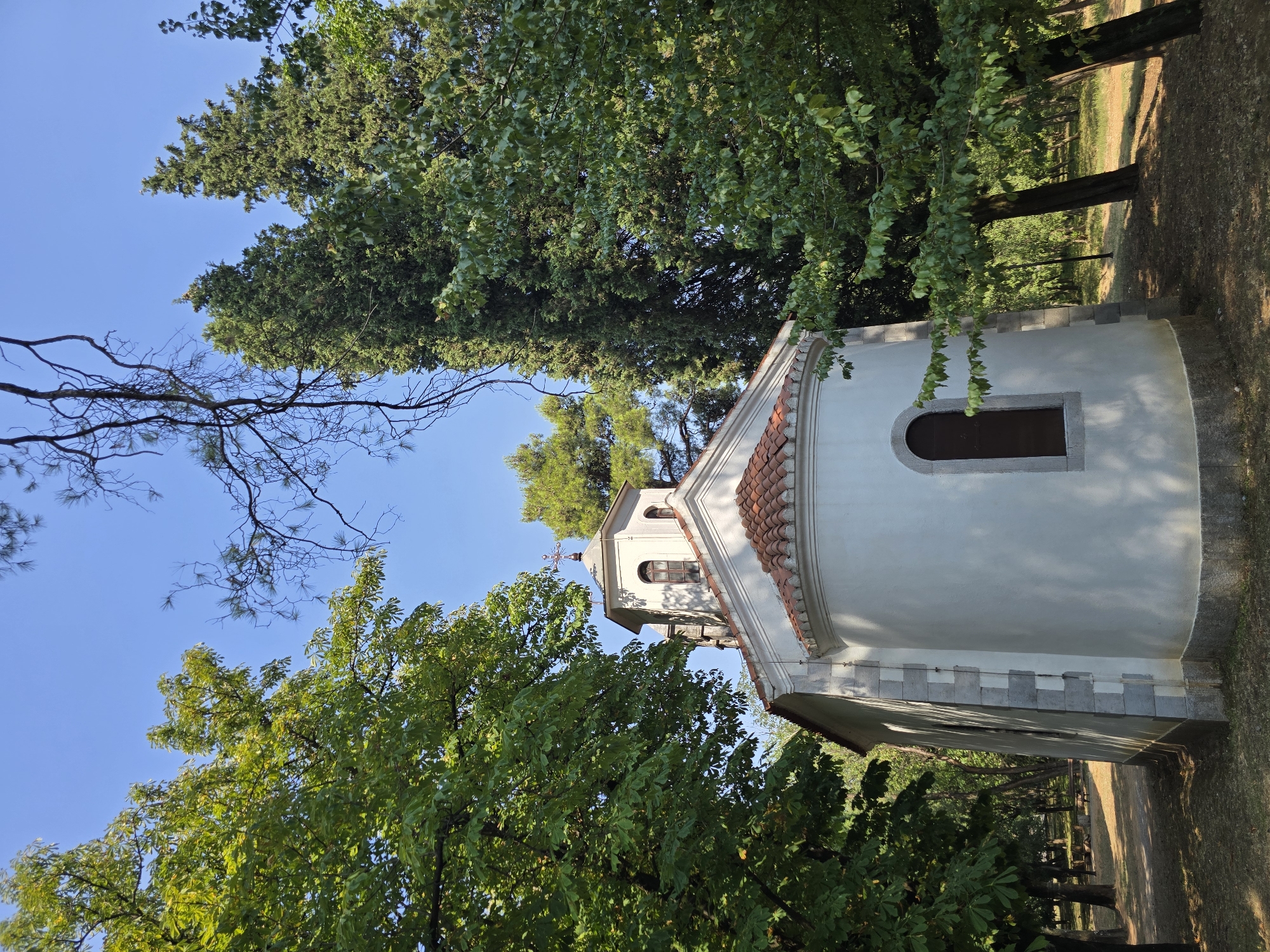 The image depicts a small, white church with a red-tiled roof, set amidst lush green trees. The church has a dome with a cross on top and arched windows. The surrounding environment appears to be a park or garden, with a dirt path and dry grass on the ground. The sky is clear and blue, suggesting a sunny day. There are no people or animals visible in the image.