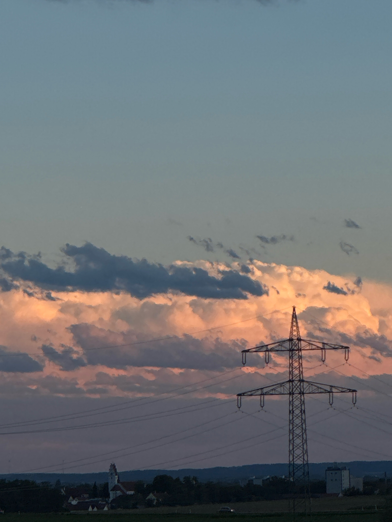 This image captures a sunset over the town of Wettstetten, Germany. A tall, lattice-style electricity pylon dominates the right side of the frame, its silhouette stark against the sky. Power lines stretch across the mid-ground, connecting to the pylon and extending out of view. In the background, the faint outline of the town of Wettstetten is visible, with a church steeple and several buildings. The sky is a gradient of soft blues and oranges, with dramatic clouds illuminated by the setting sun, casting pink and peach hues. A few scattered trees and a distant, low-rise building can also be seen on the horizon. A car is driving on a road in the lower left portion of the frame. The overall scene is one of natural beauty juxtaposed with man-made infrastructure.