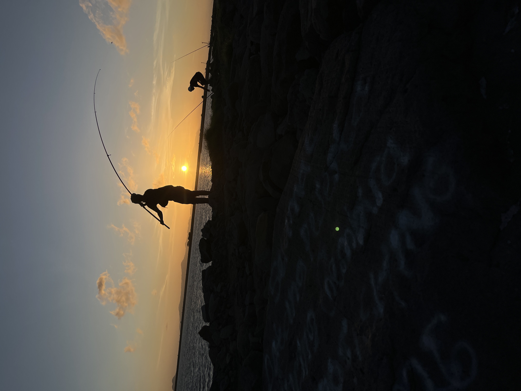 This image depicts two individuals fishing on a rocky shoreline during sunset. The sun is low on the horizon, casting a warm orange glow across the sky and reflecting on the calm water. The silhouettes of the two fishermen are clearly visible against the bright sky. The fisherman in the foreground is standing, holding a fishing rod that is bent, suggesting a catch or significant tension. The second fisherman, further in the background, is crouched, also with a fishing rod. The shoreline is composed of dark, jagged rocks, with some vegetation visible in the distance. The overall scene evokes a sense of tranquility and a typical evening pastime in a coastal area. The image quality is somewhat grainy, likely due to low light conditions and possibly a mobile phone camera. The foreground rocks have some white markings, possibly graffiti or natural patterns. There is a bright green speck visible in the lower center of the frame, likely lens flare or a light artifact. Without further information, it is not possible to definitively place this scene within the city of Banda Aceh, Indonesia, although the coastal setting and sunset are consistent with many locations there.