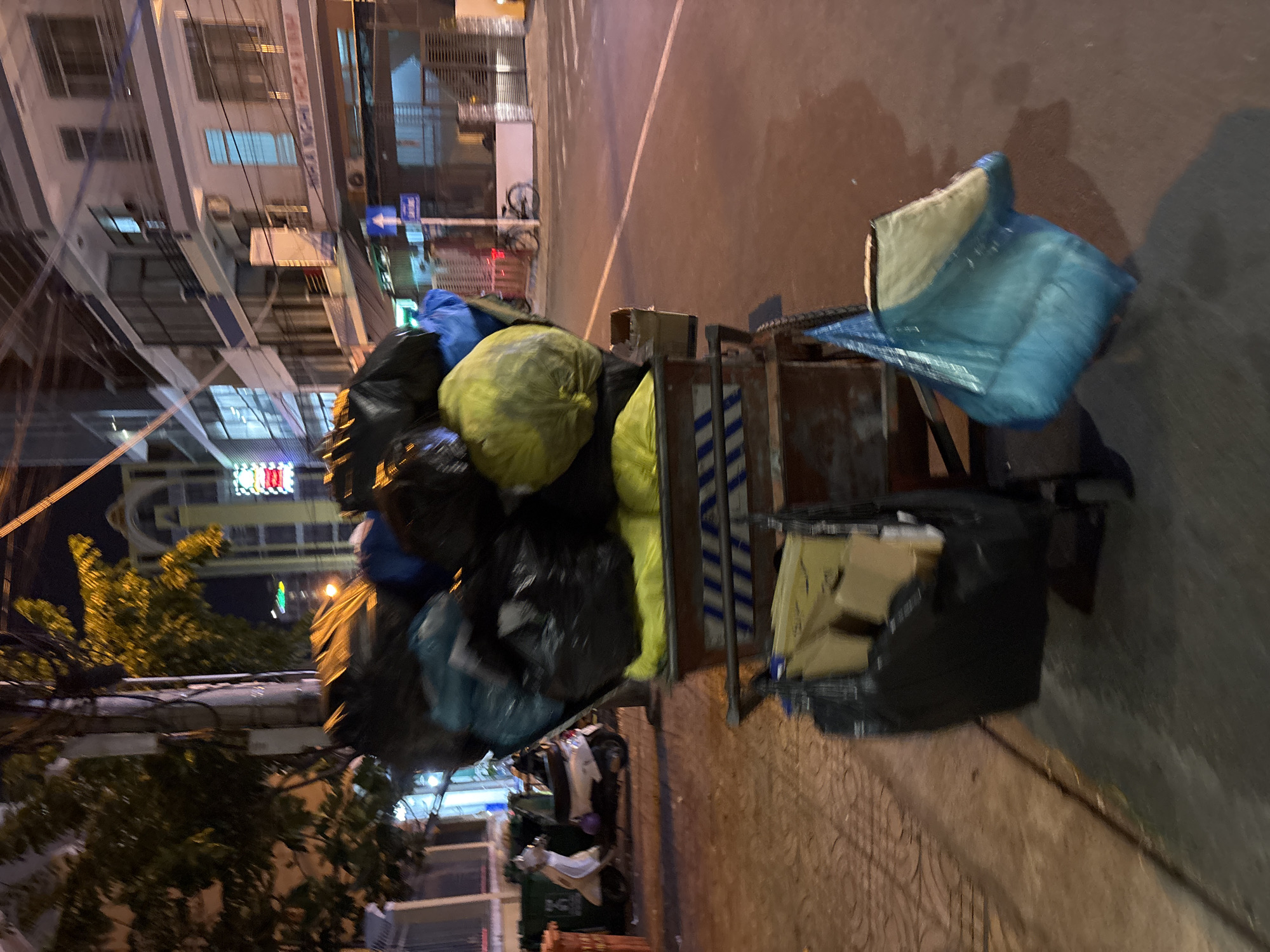 The image depicts a street scene at night in Nha Trang, Vietnam, likely a residential or commercial area. A large, overloaded trash cart is the central subject, filled with numerous black and yellow garbage bags, as well as some loose items. The cart is positioned on a paved sidewalk or street. In the background, there are multi-story buildings with lit windows, suggesting an urban environment. A motorcycle is parked on the left side of the image, and a bicycle is visible further down the street on the right. The street appears to be relatively empty, with no people prominently featured in the immediate vicinity of the trash cart. The lighting suggests nighttime, with streetlights and building illumination casting shadows and reflections. The overall impression is of a typical urban night scene with municipal services preparing for waste collection or having recently passed.