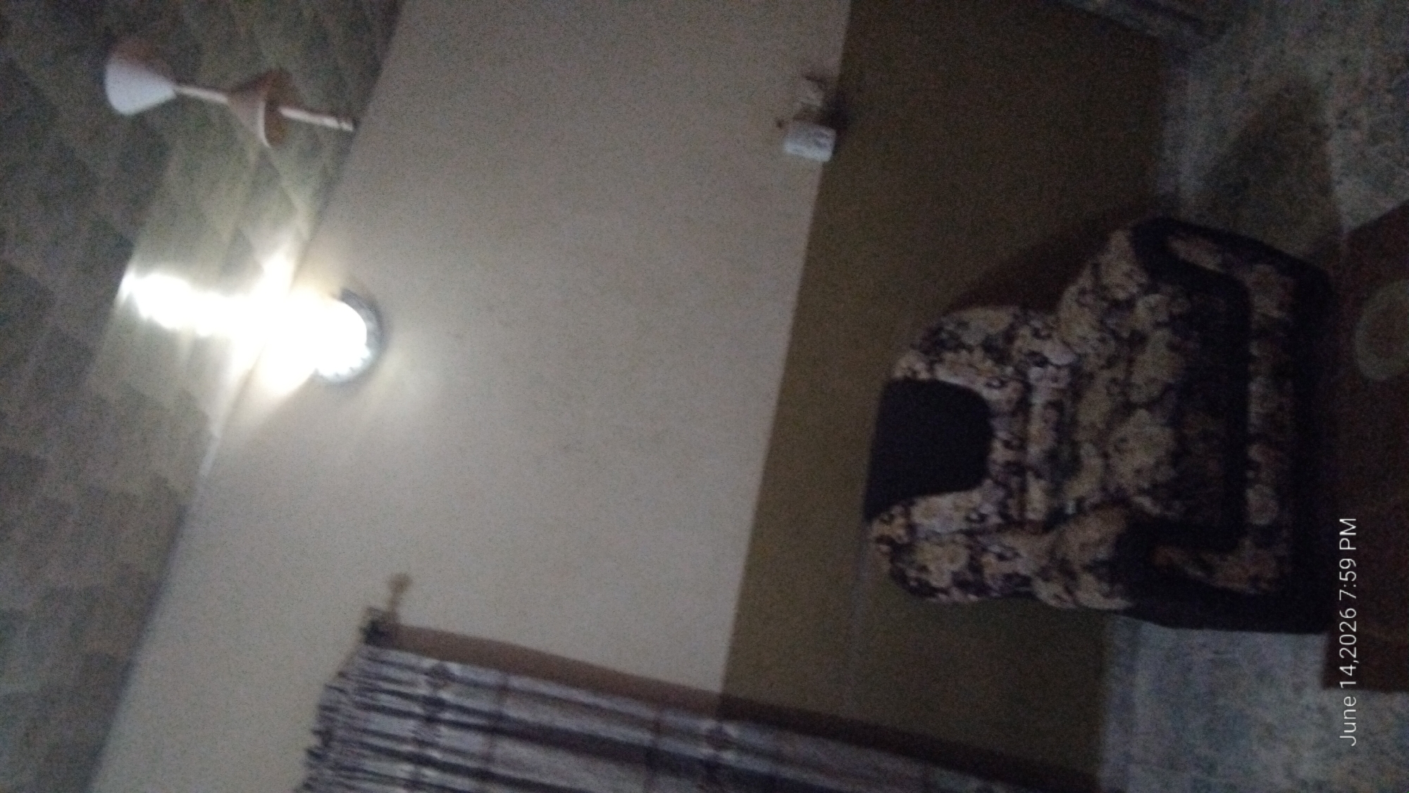 The image depicts an interior scene, likely a living room, with a patterned armchair as the central subject. The walls are painted in two tones, with a lighter shade on the upper portion and a darker brown on the lower half. A ceiling fan is visible at the top right, and a bright light fixture illuminates the center of the ceiling. On the left, a curtain with vertical stripes hangs partially drawn. The floor appears to be tiled or carpeted with a speckled pattern. A small white object, possibly a dispenser, is attached to the wall on the right. The timestamp "June 14, 2026 7:59 PM" is visible in the bottom left corner. The image is blurry and grainy, suggesting it was taken in low light conditions or with a low-resolution camera. There are no people present in the image. The location is identified as Howayi, Nigeria.