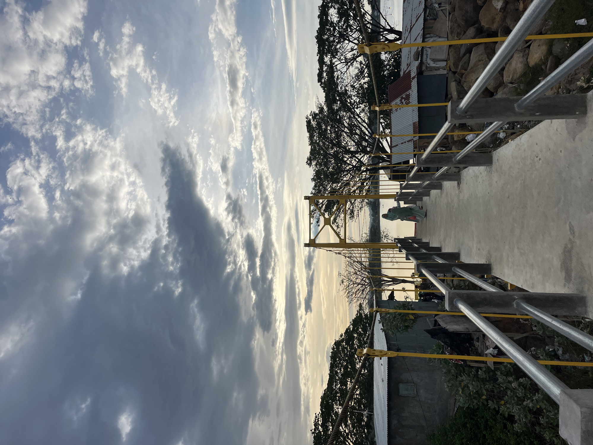 The image depicts a concrete walkway with metal railings, likely a bridge or elevated path, in Lhokseumawe, Indonesia. The sky is filled with clouds, ranging from light and wispy to dark and dense. Vegetation and rudimentary structures are visible on either side of the walkway. A person stands on the path, wearing a green garment. The scene is captured in a slightly angled, horizontal orientation.
