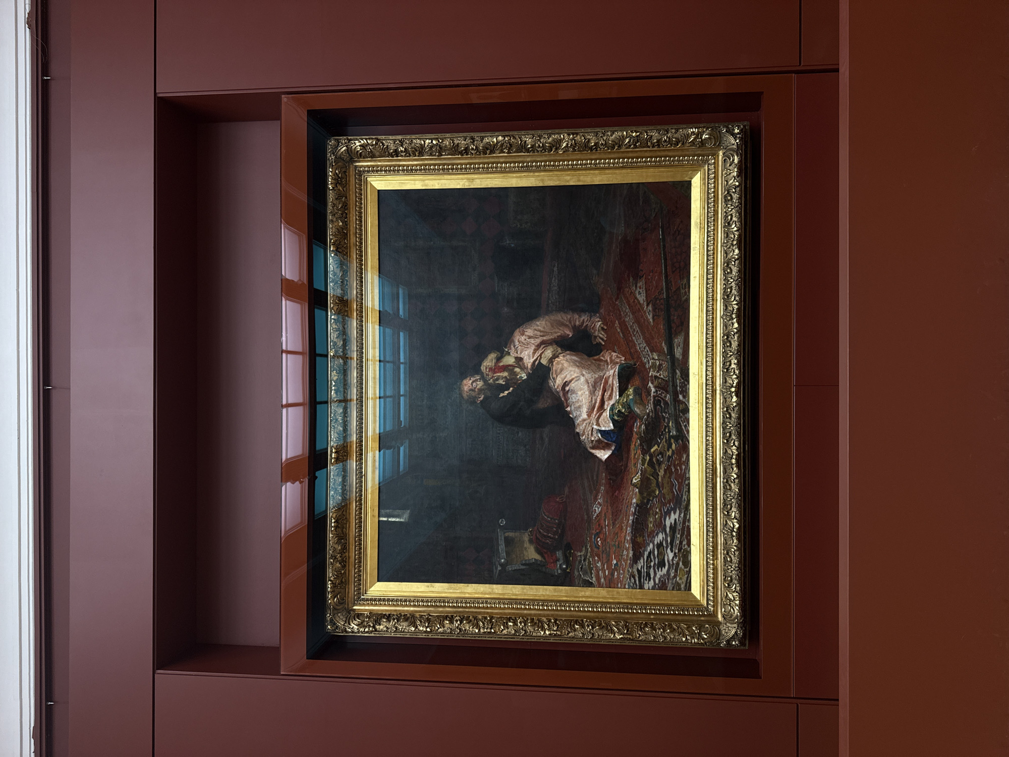 A framed painting hangs within a recessed display on a wall. The wall is composed of dark red panels. The painting features a golden frame and depicts a scene of figures in what appears to be an interior. Light reflects off the surface of the painting, obscuring some details. The painting is hung in the Tretyakov Gallery in Moscow, Russia.