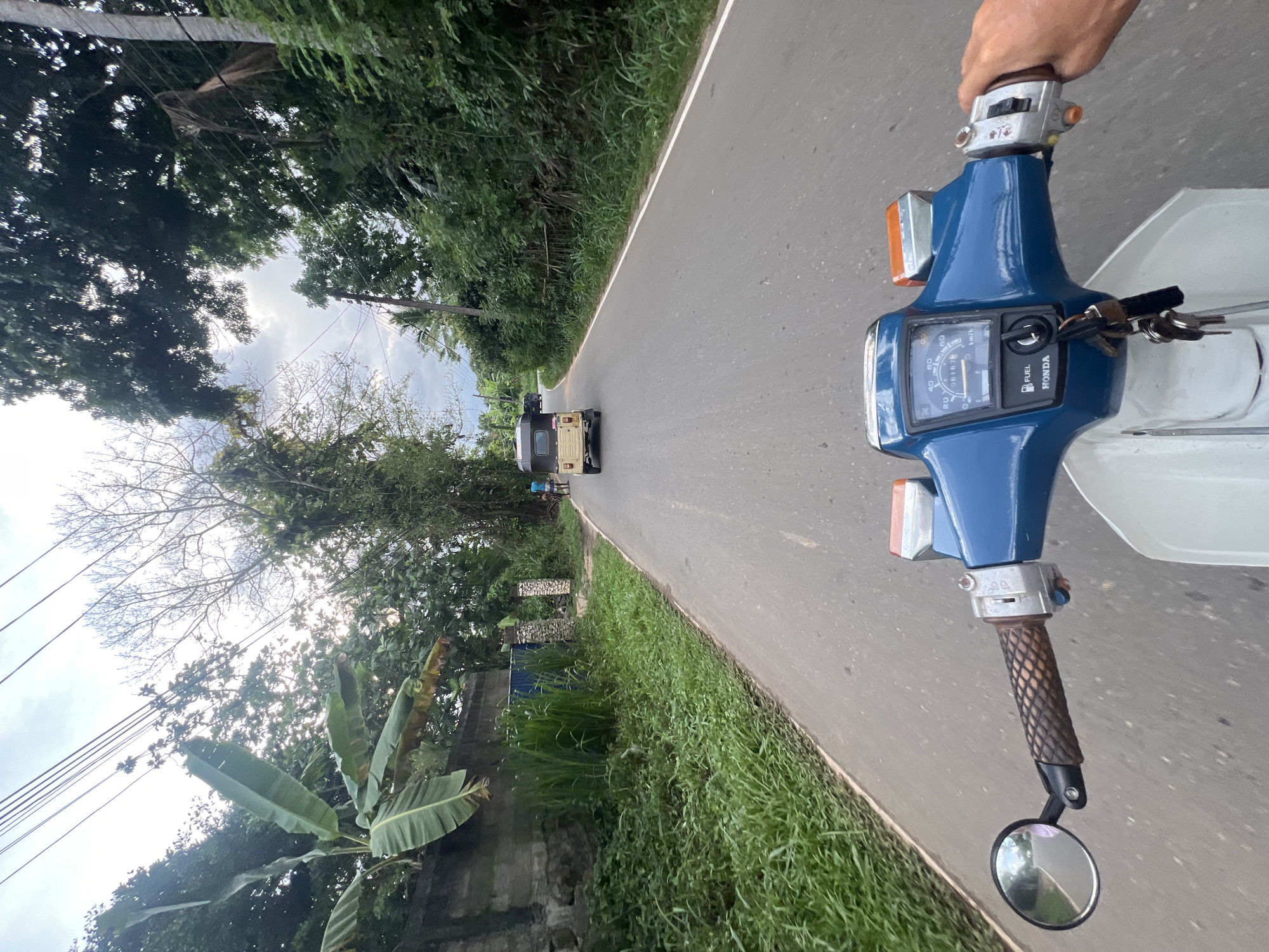 A point-of-view shot from a blue and white Honda motorcycle on a paved road outside Angulugaha Junction, Sri Lanka. A beige tuk-tuk is visible ahead in the distance. The foreground includes the motorcycle's handlebars and a partial view of the driver's right hand. Lush green vegetation borders the roadside. Overhead, trees and power lines are visible against a cloudy sky. A banana plant is visible along the lower left edge.