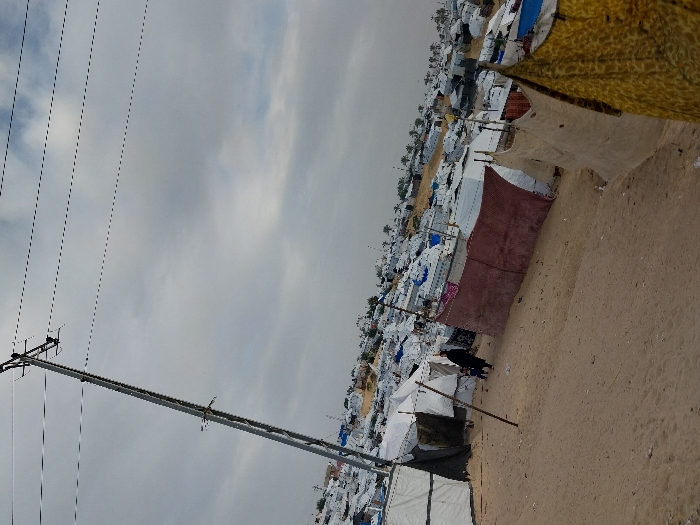 Tents and shelters cover a sandy landscape under a cloudy sky. A long line of tents stretches into the distance, creating a camp-like environment. Power lines and poles stand prominently in the foreground. The tents vary in color and construction. The overall scene suggests a temporary or makeshift settlement.