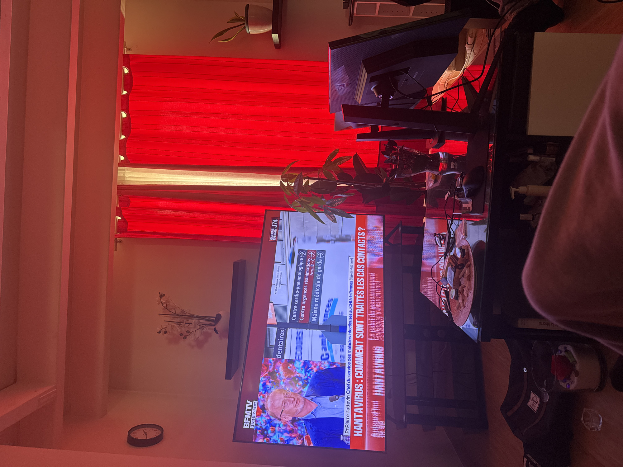 The image shows a view of an interior space, likely a room in a house or apartment. The focal point is a television screen displaying a news program from BFM TV, a French news channel. The broadcast is about the Hantavirus and features Pr Pierre Tattevin from the CHU de Rennes. Text on the screen provides information about medical services and locations, including "Centre cardio-pneumologique" and "Maison médicale de garde." The news ticker indicates "GUERRE EN IRAK J74," likely referring to developments regarding Iraq.
In the background, red and white curtains obscure the window. There are plants placed on or around a table. Other objects include a computer monitor and various items on the table like a pizza plate.
The image does not provide sufficient information to determine a specific location within the city of Paris or precise details about the people or their actions other than watching television.