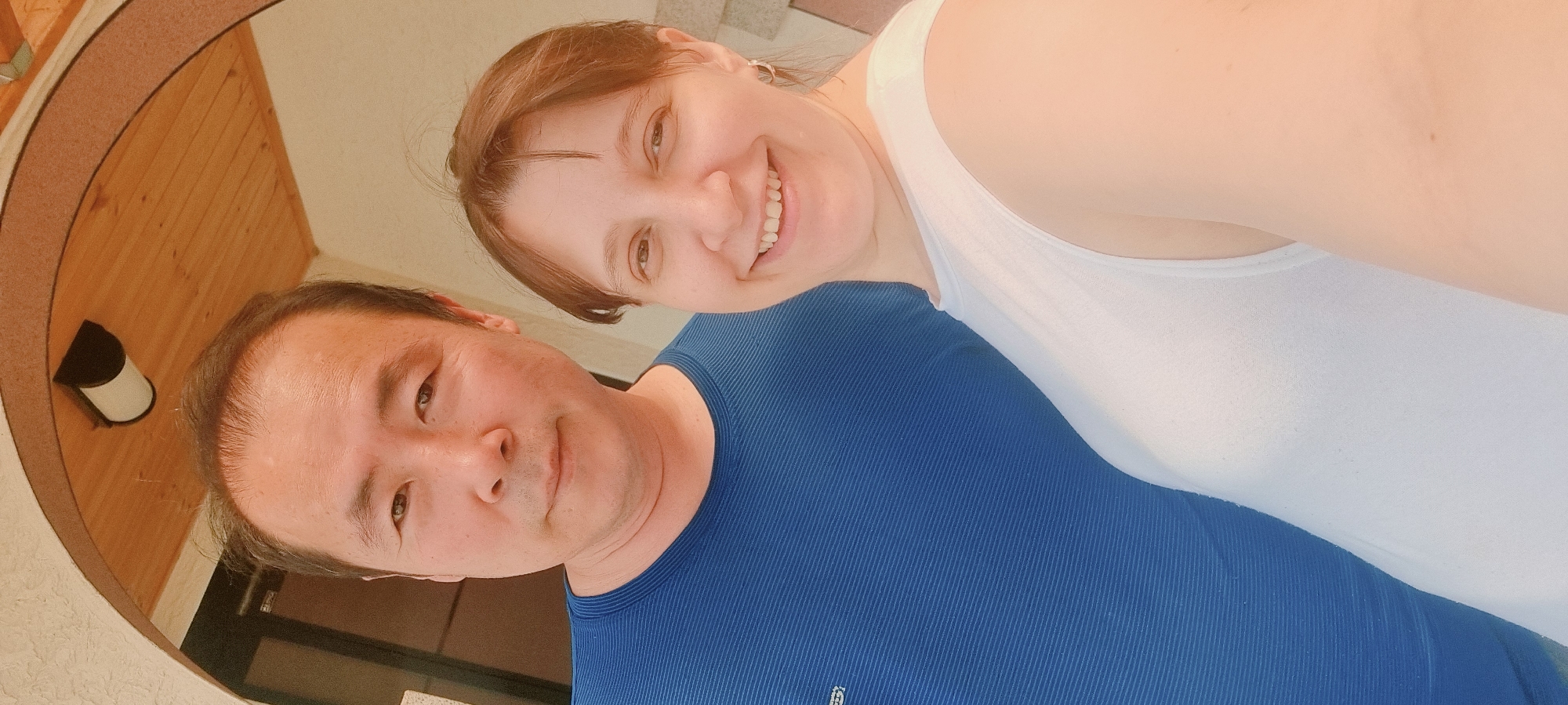 A selfie photo shows a man and a woman. The woman is smiling and wearing a white tank top. The man is wearing a blue shirt. They are standing close together. The location appears to be outdoors, possibly on a porch or patio, evidenced by the surrounding structure and outdoor lighting. The photo is taken at an angle. The exact location in Dunnae-myeon, South Korea, cannot be definitively determined from the image.