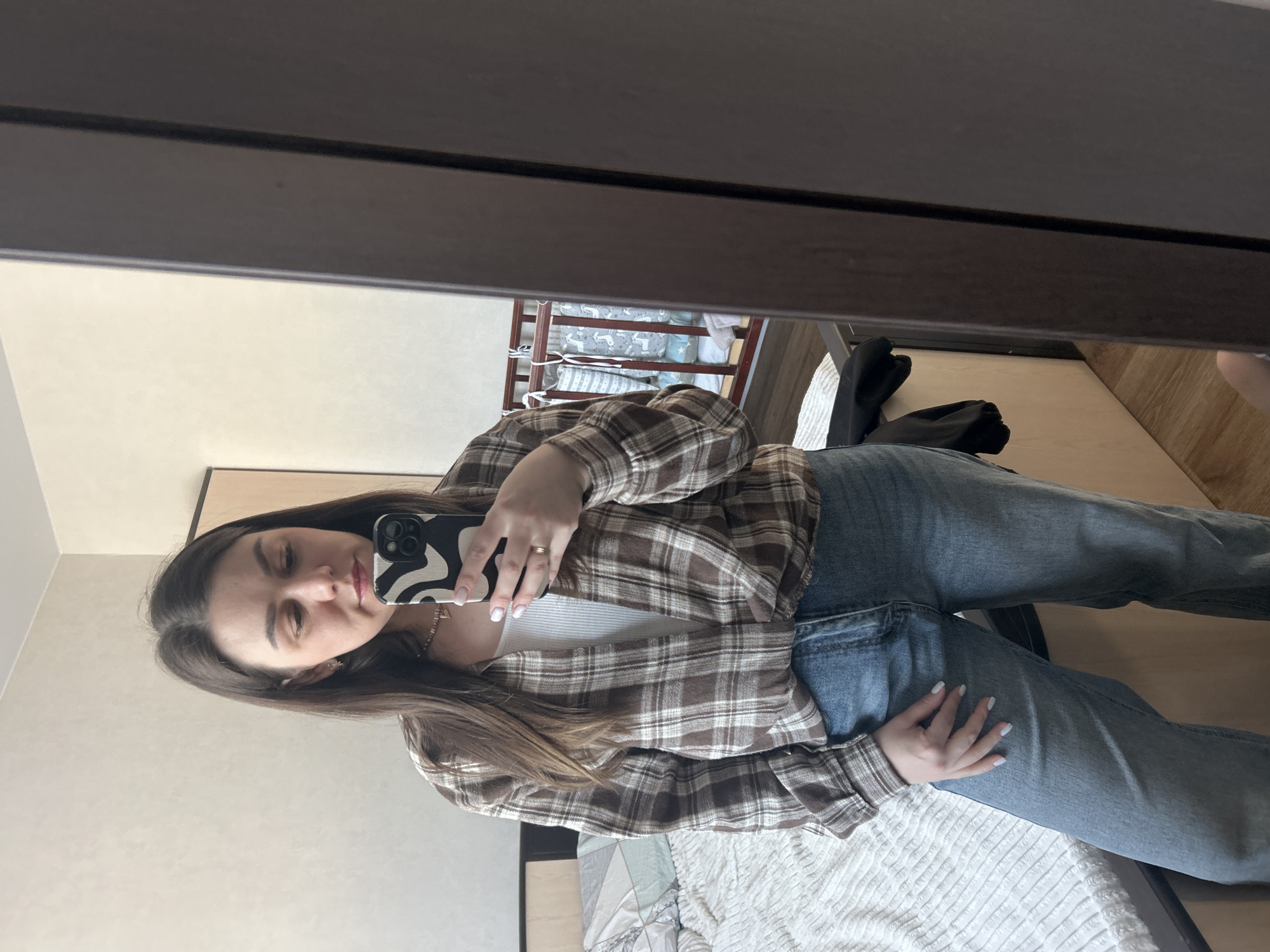 A female individual stands indoors, reflected in a full-length mirror, taking a selfie with a smartphone. The scene is located within a private residence in Chaikovsky, Russia.
The subject is a woman with long, dark brown hair, wearing a brown and white plaid flannel shirt unbuttoned over a white ribbed tank top, and light blue jeans. A gold-colored ring is visible on her left ring finger, a beaded necklace, and small star-shaped earrings are present. Her nails are manicured with light polish. She holds a smartphone with a black and white wavy patterned case in her right hand, positioned in front of her face, with her gaze directed towards the screen.
The background, visible in the mirror's reflection, depicts a bedroom setting. To the left, a bed with a light-colored wooden headboard and a darker upholstered section is present, covered with a patterned duvet and a white textured blanket. To the right, a dark brown wooden baby crib is visible, dressed with grey and white animal-themed bumpers and bedding. A white towel or blanket is draped over the crib rail. The walls are painted a light beige or cream color. The floor is covered with light wood-toned laminate or parquet flooring. A dark piece of clothing lies on a surface near the bottom right of the frame, adjacent to the bed.