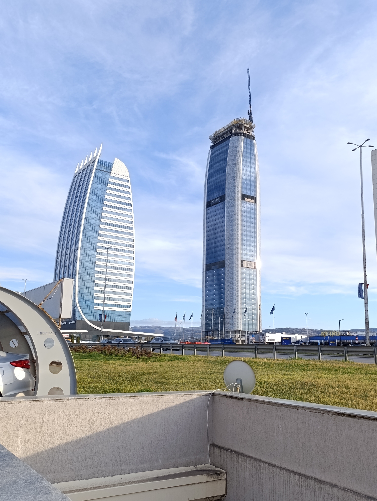 The image shows a street scene in Sofia, Bulgaria, featuring two prominent skyscrapers: the Capital Fort building (left) and the Millennium Center (under construction, right). The Millennium Center has a construction crane on top. Flags line the road behind a grassy median. The foreground includes an architectural structure, possibly part of a parking system, a satellite dish and a concrete wall. Some cars are traveling down the road. The sky is blue with some clouds. The general area is consistent with the outskirts of Sofia, near the Sofia Airport and the European Route E80. A Metro store is visible in the background.