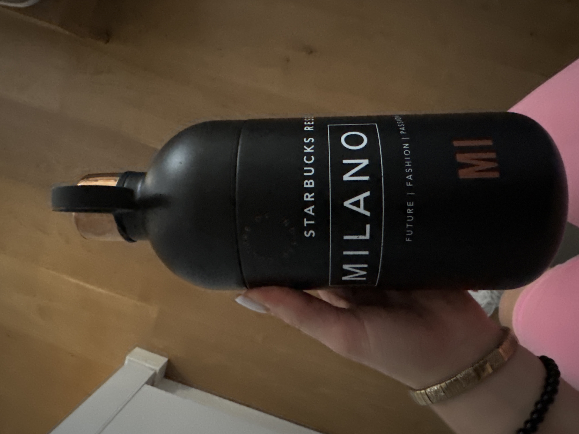 The image shows a person's hand holding a black Starbucks Reserve thermos with "MILANO" printed on it. The thermos also has "STARBUCKS RES" and "FUTURE | FASHION | PASSION" printed on it, along with "MI". The background is a wooden floor and a white baseboard. The image is taken indoors and does not depict any specific location in Murcia, Spain.
