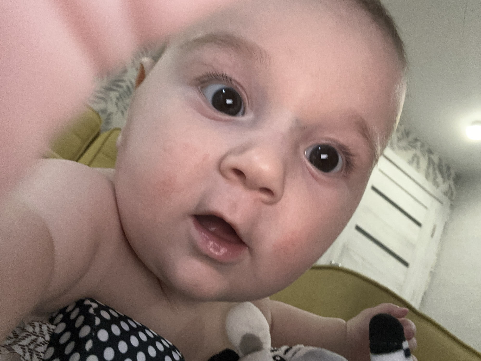 A very close-up, low-angle portrait captures an infant with wide, dark brown eyes staring directly at the camera. The baby's face fills most of the frame, showcasing fair skin with rosy cheeks and a slightly open mouth that reveals a tongue but no visible teeth, suggesting curiosity or a moment of mild surprise. Light, sparse hair is visible on the baby's head.
The baby appears bare-chested or in minimal upper body clothing. On the lower left, a black fabric with white polka dots is visible, possibly part of a blanket or garment. The baby's small hand on the lower right gently holds a soft toy with white and black elements.
The background is intentionally blurred due to the close proximity of the camera to the baby. Details discernible in the soft-focus background include a yellow or gold-colored piece of furniture, likely a sofa or armchair, on both the left and right sides. In the upper right, a white door with two distinct dark horizontal panels is visible, along with a soft light source, possibly a ceiling light. A subtle, light-colored patterned wallpaper or fabric can be seen on the wall behind the baby's head on the left.
The image conveys a tender, intimate moment, likely a "baby selfie," emphasizing the baby's innocent and inquisitive expression within a cozy, domestic setting. There is no visible text in the image.