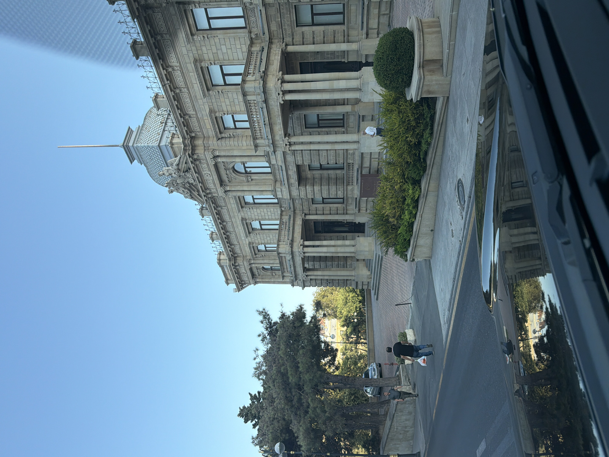 A wide shot captures the ornate facade of the Palace of Happiness (also known as the Palace of the newlyweds) in Baku, Azerbaijan, under a clear blue sky. The building, constructed of light-colored stone, features classical architectural elements such as columns, balconies, and decorative sculptures. In the foreground, a black vehicle's hood is visible, reflecting the surrounding environment and a glimpse of the street. A paved walkway leads to the palace entrance, lined with manicured green bushes and trees. Two men are walking on the street; one is in a dark shirt carrying a white bag, while the other, in a lighter shirt, walks ahead. A silver car is parked on the side of the road. The overall scene suggests a bright, late afternoon in the city.