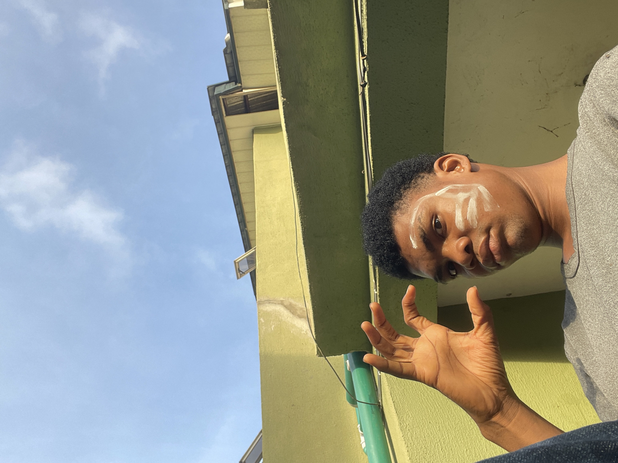 A low-angle medium shot shows a person outdoors next to a building. The person has dark skin, short black hair, and white markings on their forehead. They are wearing a gray shirt and blue jeans. Their right hand is raised with fingers extended. The building is pale green with a vertical green pipe. The sky above is blue with scattered clouds. The location may be Ado, Nigeria based on the prompt, but no specific evidence in the image confirms this.