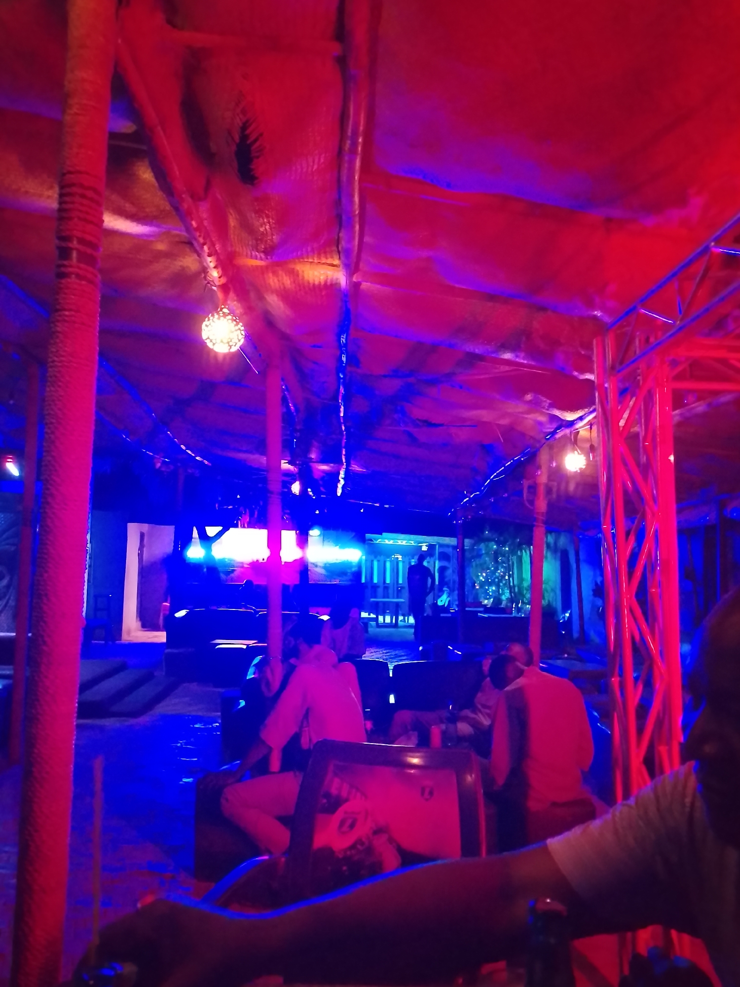 The image depicts an indoor event space in Unknown, Unknown, with a red and blue lighting scheme. A textured, tent-like roof structure is present, supported by several red columns. There are people seated at tables and lounge areas, with a stage area in the background. A man is visible in the foreground, possibly seated at a bar or table. The overall atmosphere appears to be festive and lively.