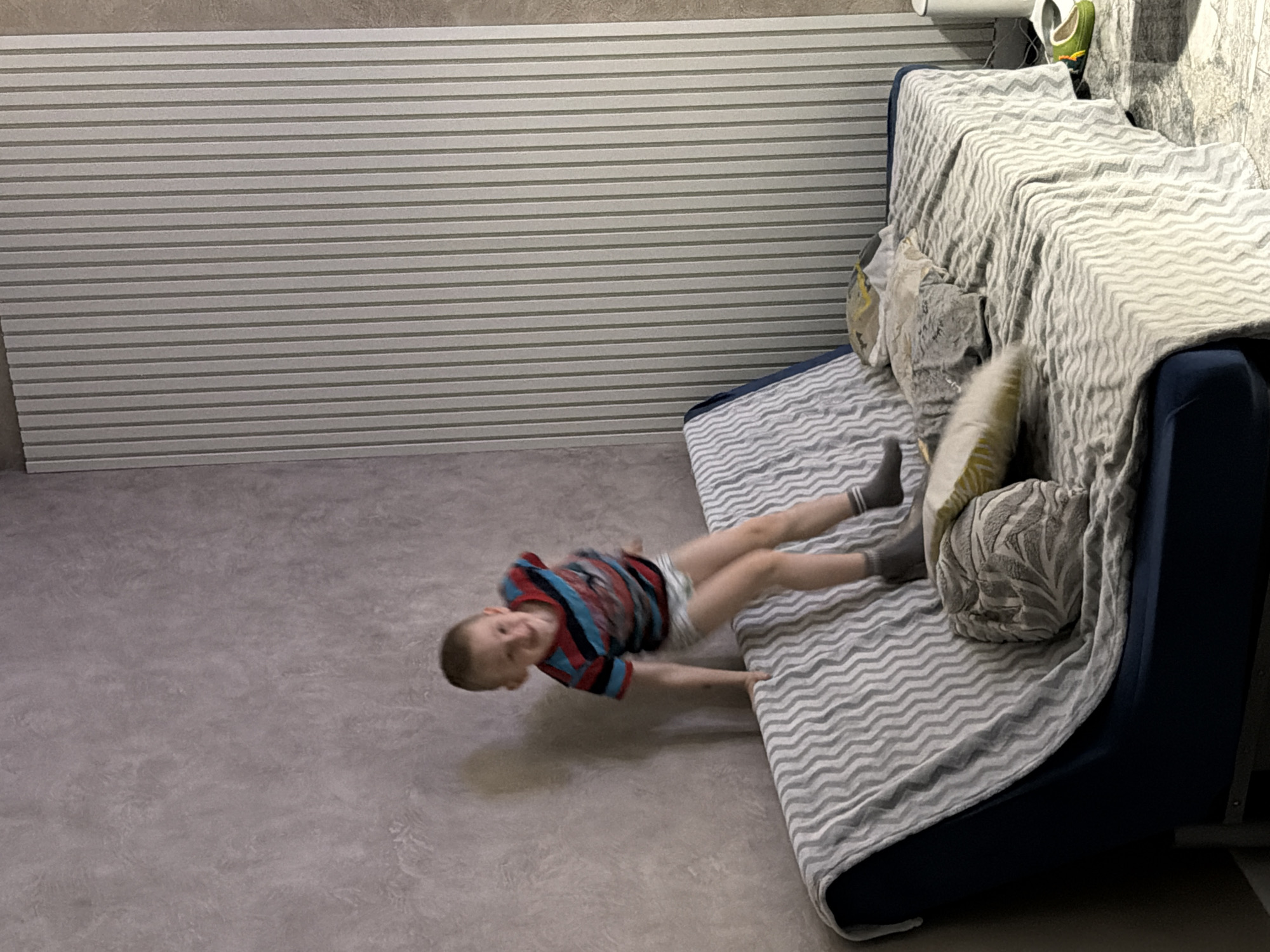 A young boy lies on a sofa positioned against a wall with horizontal striped wallpaper. He is wearing a short-sleeved, red and blue striped shirt, light-colored shorts, and grey socks. The sofa has a grey and white zig-zag patterned cover and several pillows. The floor is a mottled grey color. The location is unspecified and there is no evidence of the city of Rossiyskiy, Russia.
