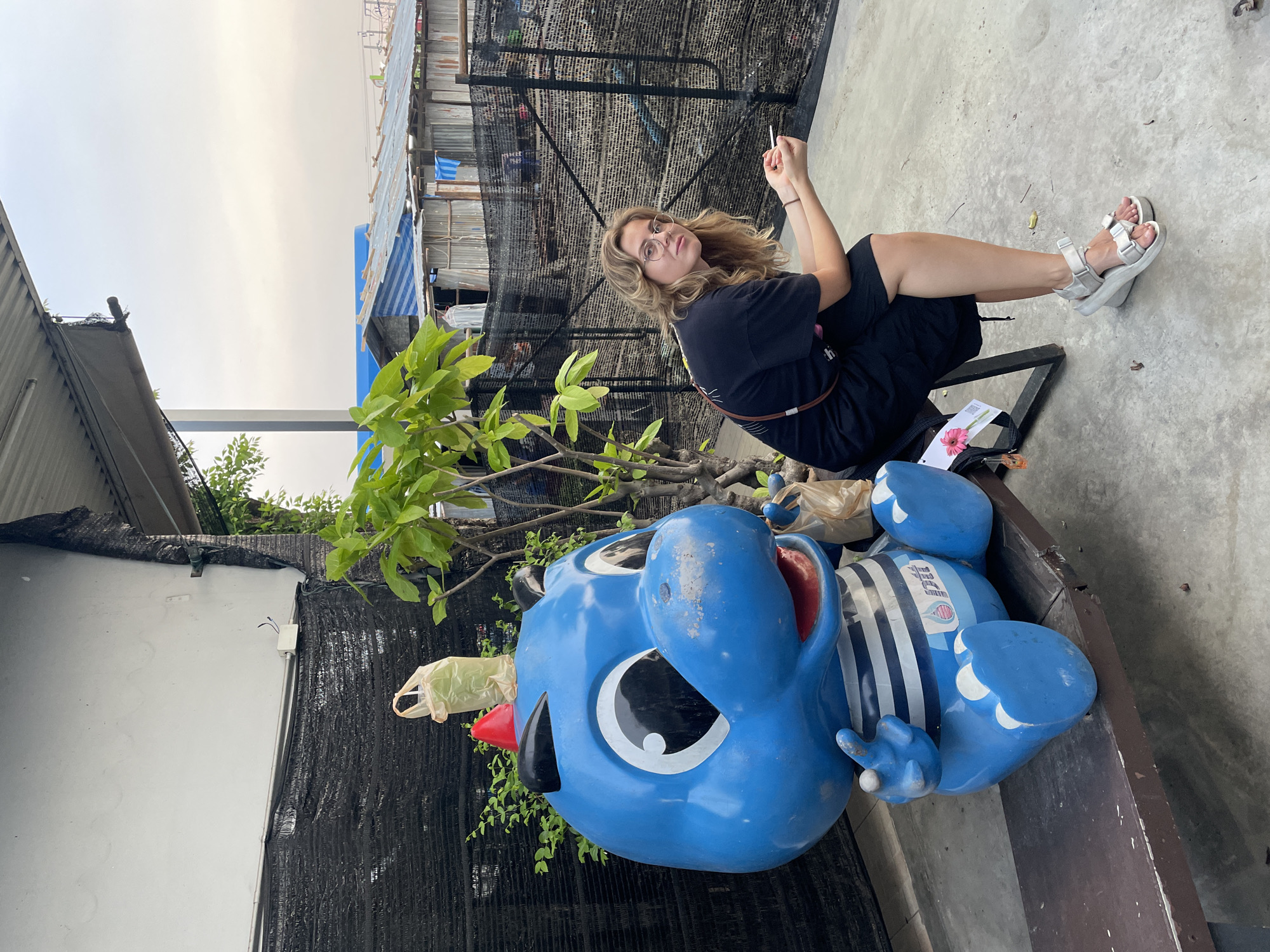 The image captures a casual outdoor scene featuring a young woman seated next to a large, blue cartoon statue, set against an urban backdrop.
The main subject, a woman with wavy blonde hair and round glasses, is positioned towards the right of the frame. She wears a black t-shirt, dark shorts, and light grey platform sandals. She sits with her legs extended, holding a small, white, stick-like object (possibly a cigarette or vape pen) between the fingers of her left hand, which is raised near her face. She looks directly at the camera with a neutral, slightly pensive expression. A brown strap, likely from a bag, crosses her chest.
To the woman's left, prominently in the foreground, rests a large, blue, cartoonish statue, lying on its back on a wooden surface. This figure has big white and black eyes, a red cone-shaped nose, and black lips. It wears a white and blue striped top with an indistinct logo on its chest. A light-colored plastic bag is caught on its red nose.
The background reveals a dense, urban environment with structures made of corrugated metal sheets and tarpaulins, indicative of informal settlements or less developed urban areas. A tall black mesh net or tarp is stretched vertically behind the woman, partially obscuring the background buildings. Green leafy plants are visible in the foreground near the statue, as well as in the upper background. The ground is concrete. The sky overhead is a uniform light grey, suggesting an overcast day. On the bench next to the woman, a piece of paper with a pink flower graphic and unreadable text is visible.
The scene conveys a relaxed, everyday moment within an urban setting, likely in Bangkok, Thailand, given the architectural style of the background structures.