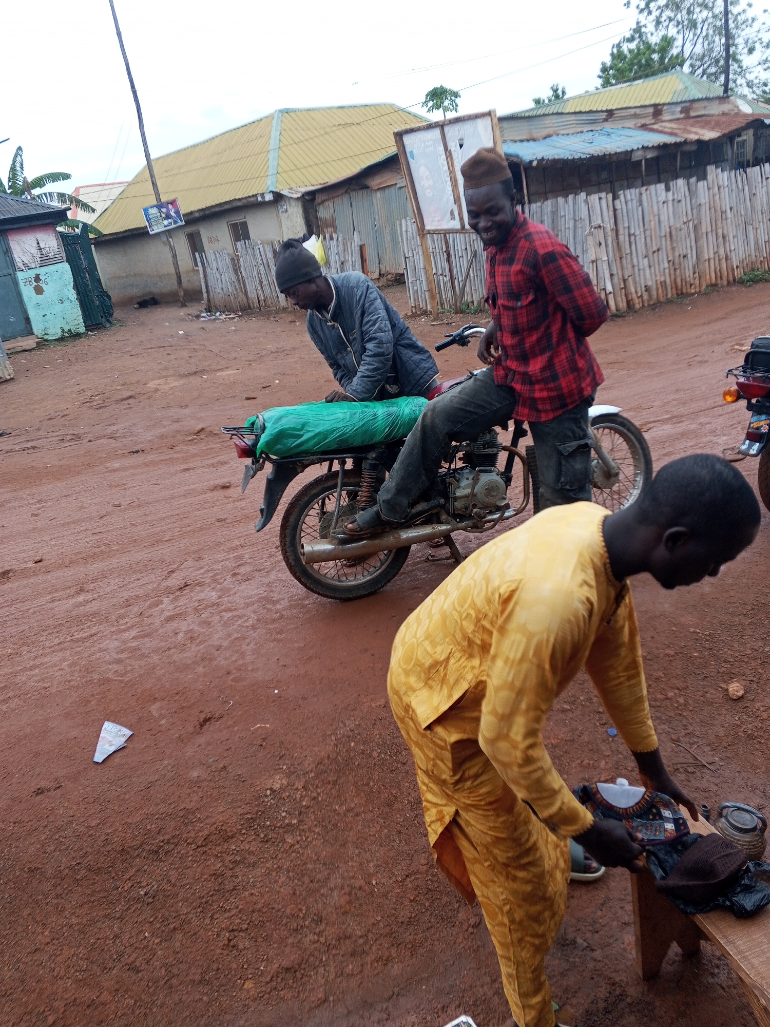 In Abuja, Nigeria, a man in a yellow traditional outfit is bent over a wooden bench. On the bench are a dark-colored cap, a small black pot, and a patterned cloth being handled by the man. Beside him, another man on a motorcycle wears a red and black checkered shirt and a brown cap. He is smiling and looking towards the viewer. The motorcycle is covered with a green tarp. A third man, wearing a dark jacket and a black beanie, sits on the motorcycle behind the man in the checkered shirt. The ground is a reddish-brown dirt road, with scattered debris including a white folded paper. In the background, there are low-rise buildings with corrugated metal roofs and bamboo fencing. A utility pole with a poster on it is visible to the left. The sky is overcast.