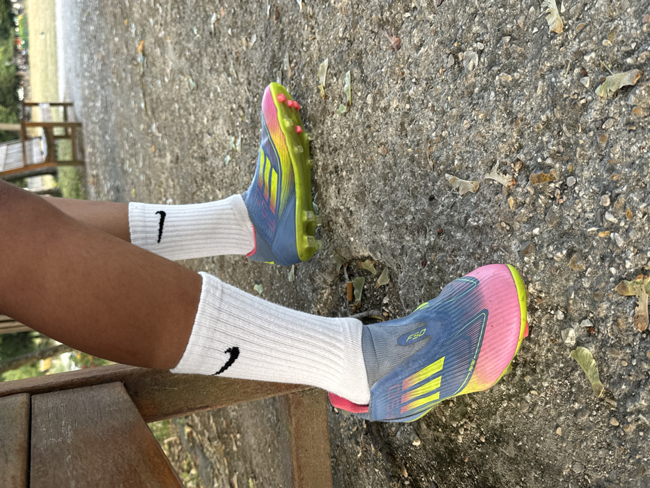 The image depicts the lower legs and feet of an individual with a dark skin tone, situated in what appears to be a park or recreational area in Conflans-Sainte-Honorine, France. The subject wears white, ribbed, crew-length Nike socks, each featuring a black swoosh logo on the outer ankle. They are shod in multi-colored Adidas F50 soccer cleats, characterized by blue, neon yellow, and bright pink hues, with neon yellow/green studded soles and pink conical studs. The left cleat exhibits a slightly looser fit around the ankle, revealing interior pink material. A portion of a weathered wooden bench is visible on the left side, partially obscuring the subject's left leg. The ground surface is a coarse, light gray, granular texture, interspersed with small stones and scattered dry leaves. A cigarette butt is present on the ground near the left cleat. The background is blurred, showing green foliage and another distant wooden bench, indicative of an outdoor, natural environment.