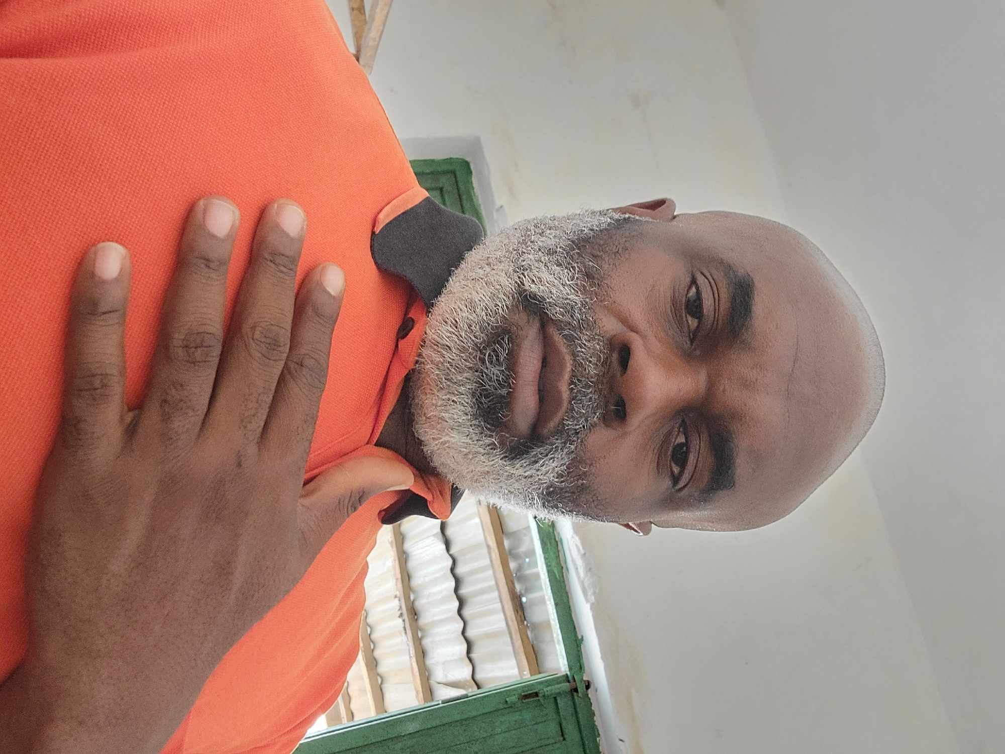A bald-headed, dark-skinned man with a gray beard is looking directly at the camera. He is wearing an orange shirt with a black collar and has his hand placed on his chest. The background shows a green-painted structure with corrugated metal visible behind it and a white wall. The image was likely taken indoors. There is no information available to determine the location is in Maiburiji, Nigeria.