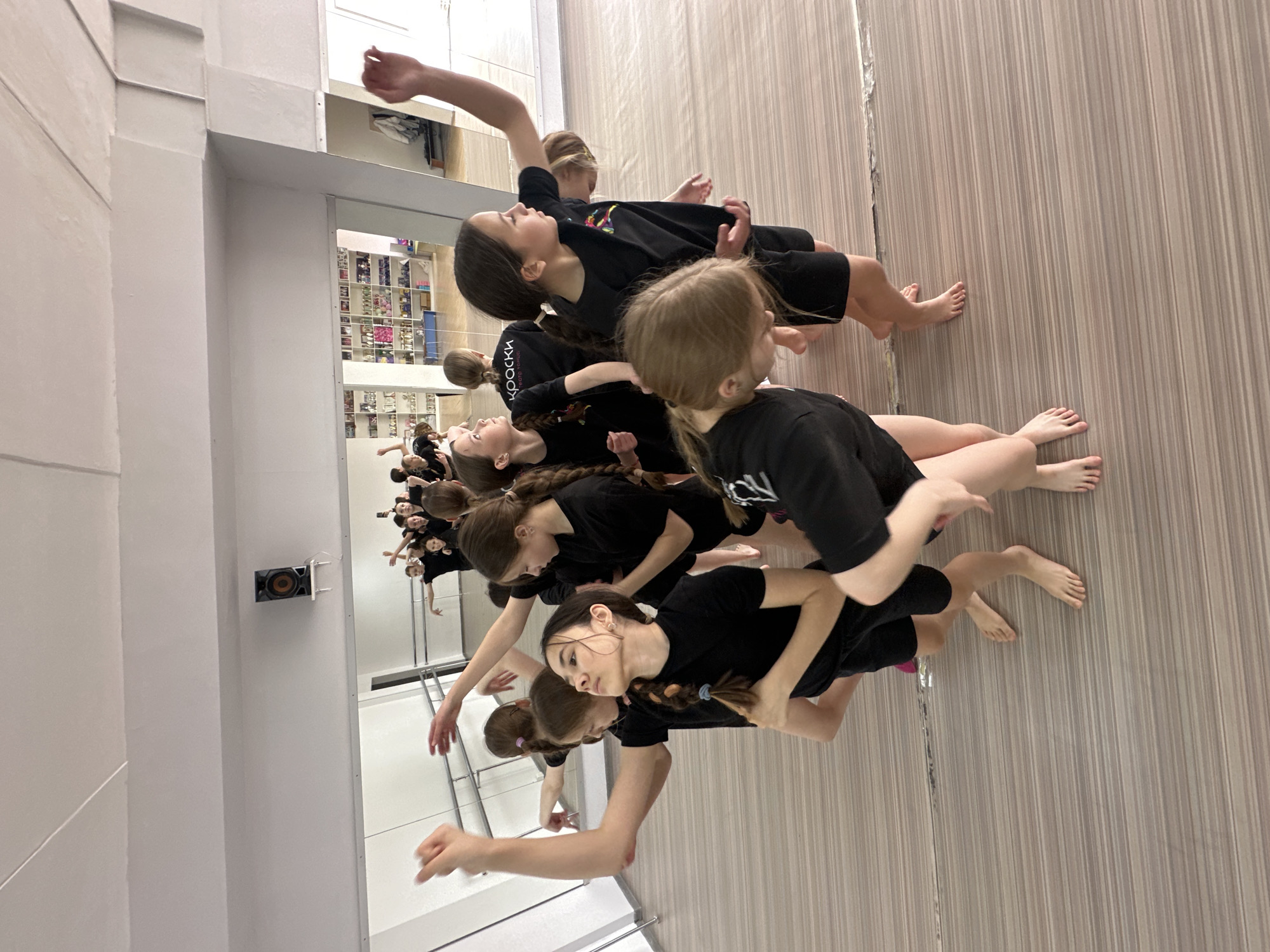 A group of young girls, likely pre-teens or early teens, are captured mid-movement in what appears to be a dance studio in Votkinsk, Russia. The scene is dominated by a large mirrored wall, which reflects the girls and a room beyond filled with shelves holding various items, possibly shoes or supplies. The girls are dressed uniformly in black t-shirts and dark shorts, with many being barefoot. They display various poses, some with arms outstretched and bodies leaning, suggesting they are performing a choreographed dance routine or engaging in a physical warm-up. Their expressions indicate focus and concentration.
On one girl's black t-shirt, the Russian word "КРАСКИ" (KRASKI), meaning "PAINTS" or "COLORS," is partially visible. The floor is light-colored, featuring a subtle pattern of vertical stripes, while the visible walls are white. A small, dark speaker is mounted on a white wall segment adjacent to the mirrored surface. The lighting is bright and even, typical of an indoor practice space. The overall impression is one of dedicated rehearsal or training for a performance.