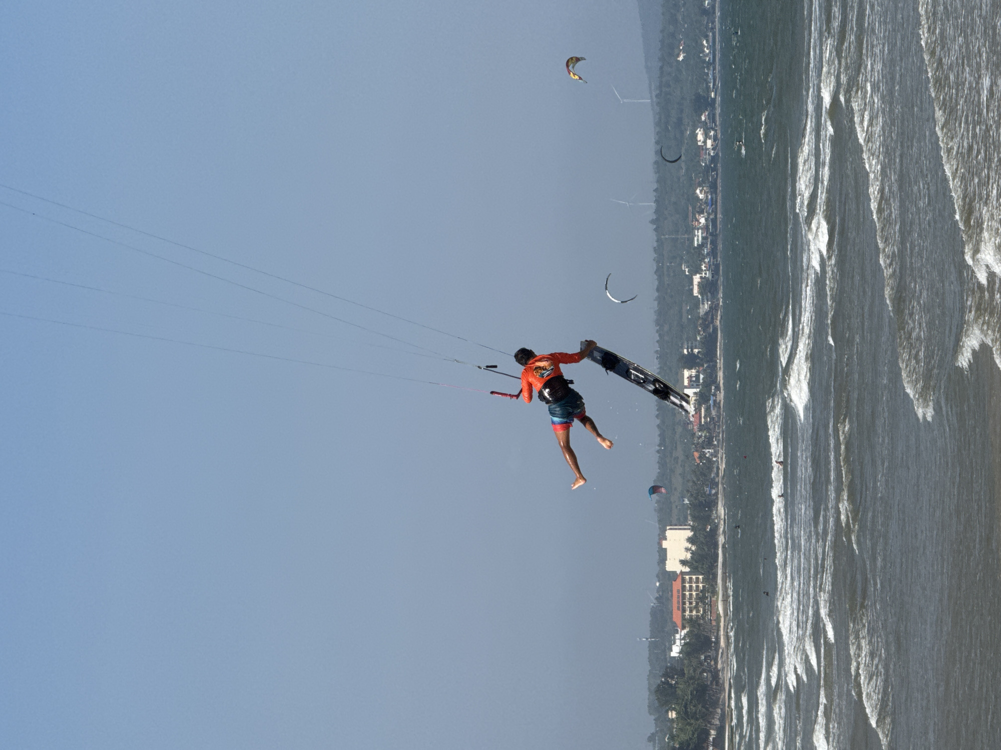 In Mũi Né, Vietnam, a kitesurfer is captured mid-air, performing a jump over the choppy ocean. The kitesurfer, wearing an orange rash guard and blue shorts, is suspended above their board, which is angled downwards. The kite, connected by lines, is taut and high in the clear blue sky. In the background, the coastline is visible with buildings, palm trees, and hills. Several other kitesurfers and colorful kites are scattered across the water and sky, indicating a popular kitesurfing spot. The waves are white-capped, suggesting a windy day suitable for the sport. Wind turbines are also visible on the distant hills.