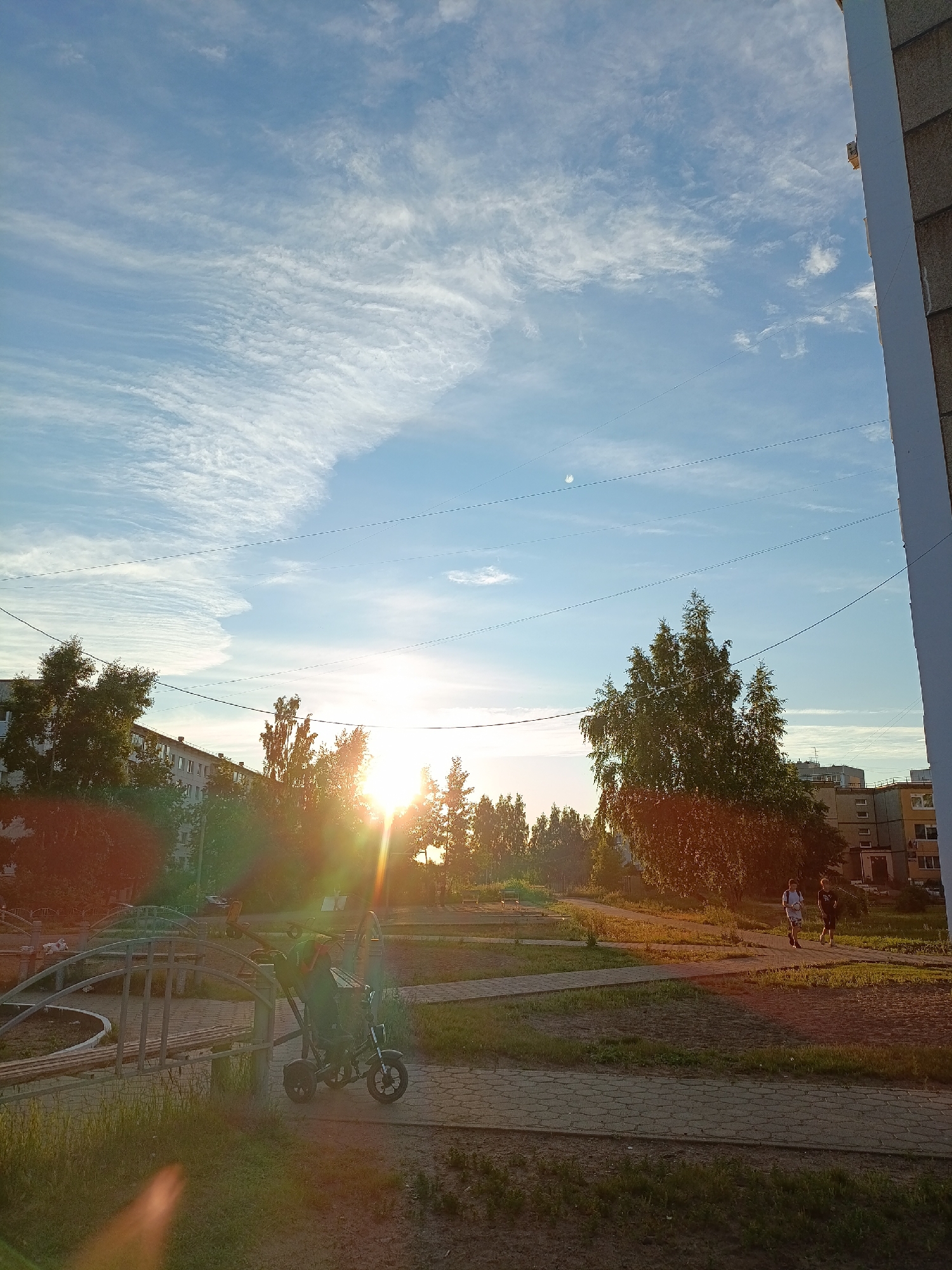 The image depicts a sunny day in Chaikovsky, Russia, likely in a residential area. The sun is low in the sky, casting a warm glow and lens flares across the scene. In the foreground, a playground area is visible, with a metal railing and bench. A child's stroller is parked near the playground equipment. Further back, two young individuals, possibly teenagers, are walking on a paved path. Trees and apartment buildings line the background, suggesting an urban or suburban setting. The sky is a bright blue with scattered white clouds. The overall impression is one of a peaceful, everyday moment in a Russian city.