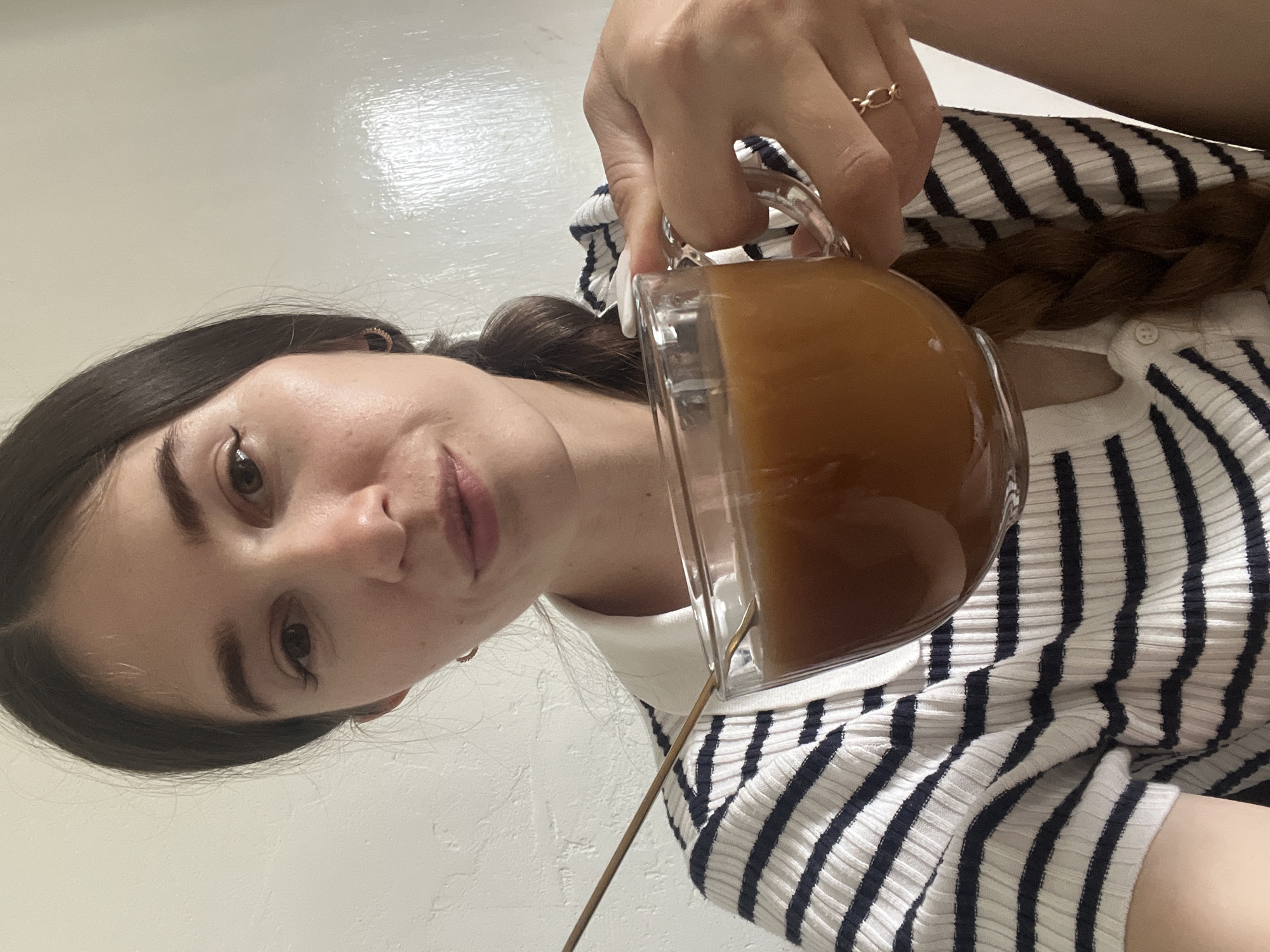 In an indoor setting with a textured white wall in the background, a young woman is shown from the chest up, taking a selfie while holding a clear glass mug filled with a light brown beverage. She has dark brown hair styled in a braid that rests on her shoulder and is wearing a white collared shirt with horizontal navy blue stripes. Her right hand is holding the mug, and a golden spoon is visible inside. A gold ring with an interlocking loop design is worn on her right ring finger.