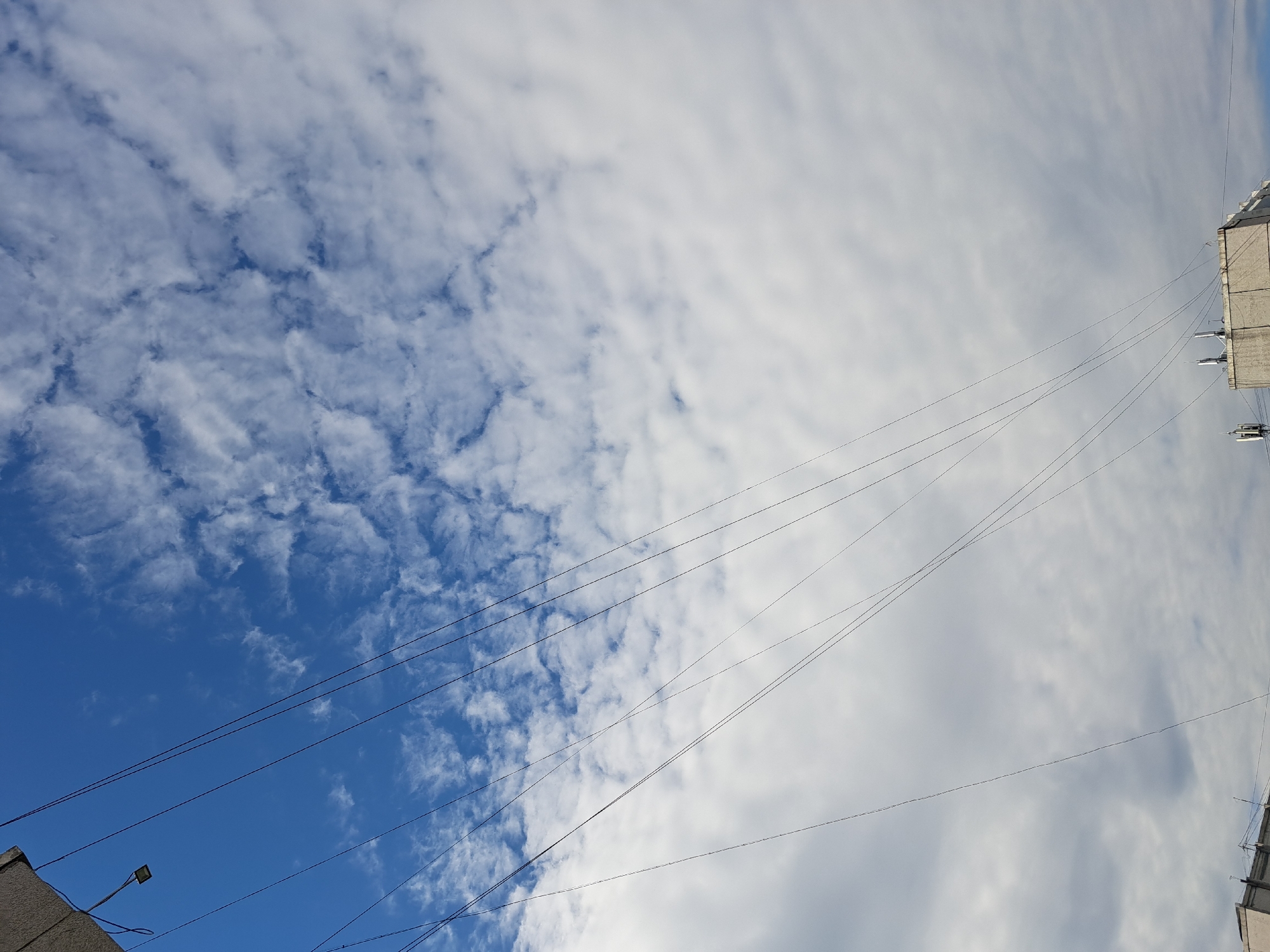 The image captures a skyward view dominated by a blue sky with scattered white clouds and a series of thin, dark wires crisscrossing diagonally from the upper left to the lower right. In the upper left corner, a portion of a building or structure is visible, with a small, rectangular light fixture attached. The lower portion of the image reveals the tops of several buildings, some with what appear to be antennae or communication equipment. The scene is consistent with an urban environment. Based on the metadata provided, the location is Irkutsk, Russia.
