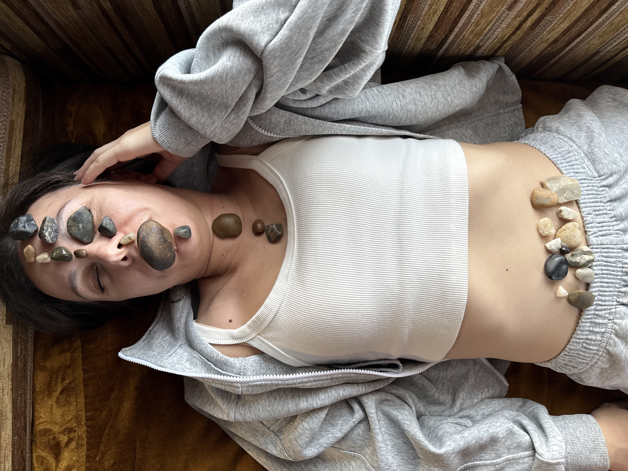 A person is lying on a striped brown couch with their eyes closed, a grey zip-up hoodie and a white tank top on. Several smooth, dark grey and light brown stones are placed on their forehead, in their mouth, on their neck, and across their lower abdomen. The person is located in Solnechnyi, Russia.