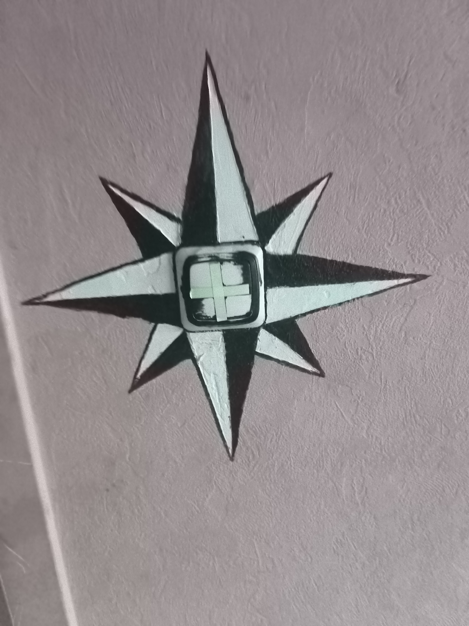 The image shows a compass rose painted on a textured, light-colored wall. A modern electronic device, resembling a smart watch, is affixed to the center of the compass rose. The watch face displays a bright, cross-shaped graphic. The lighting in the image is soft, casting subtle shadows from the painted elements and the device. The environment appears to be indoors, with a section of a wall visible in the foreground and a brighter, out-of-focus area on the left side, possibly indicating a doorway or a different illuminated section. There are no people or animals visible. The overall impression is one of decorative art integrated with modern technology. Without further information, the specific location in Riga, Latvia, cannot be determined from the visual content of the image alone.