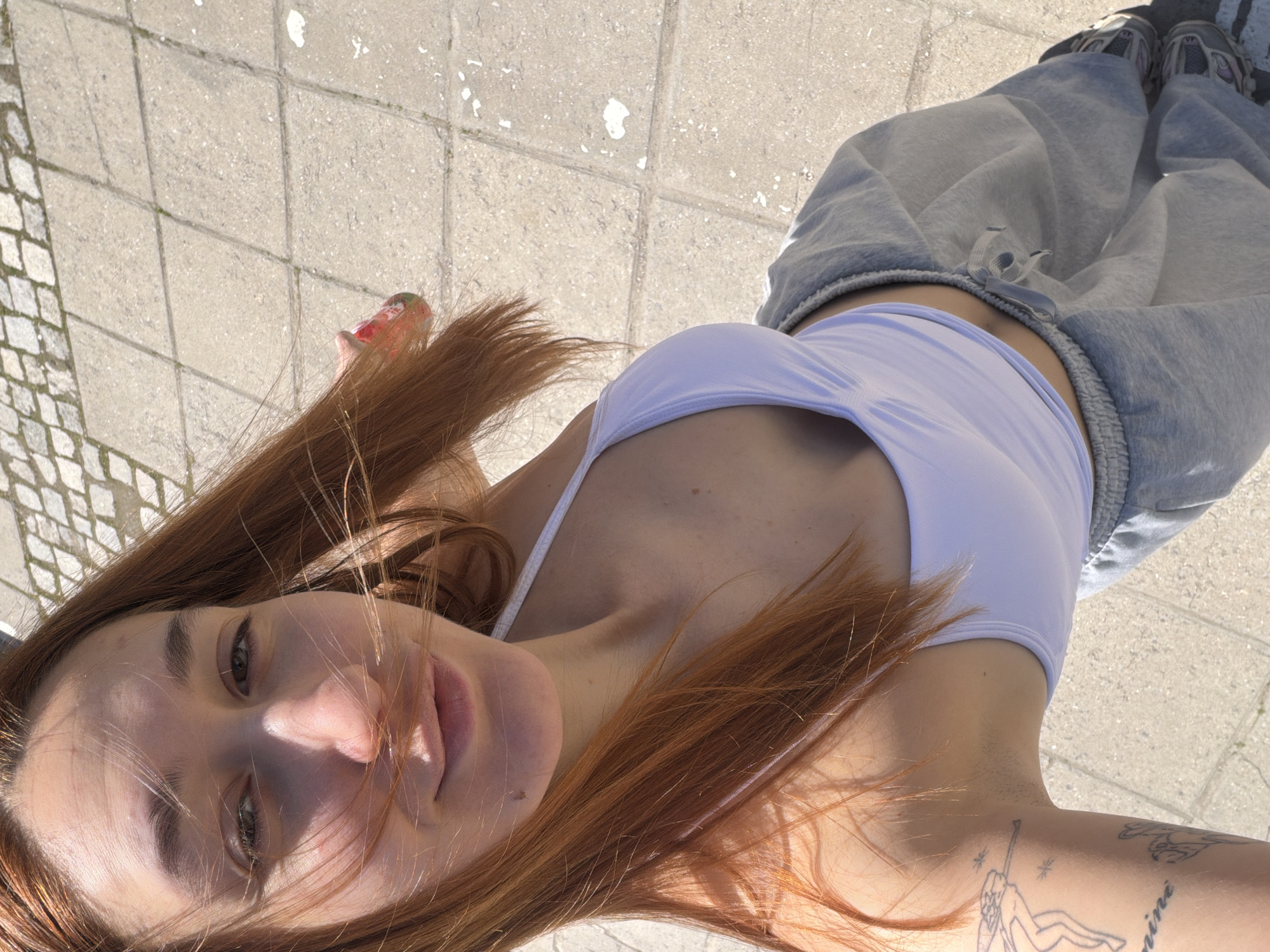 A young woman lies on her back on a paved surface, possibly a sidewalk, in an outdoor location. She is wearing a light-colored tank top and grey sweatpants. Her long, reddish-brown hair is partially obscuring her face. The paving appears to be a mix of large, rectangular tiles and smaller, cobblestone-like sections. The location is not identified within the city of Sofia, Bulgaria.