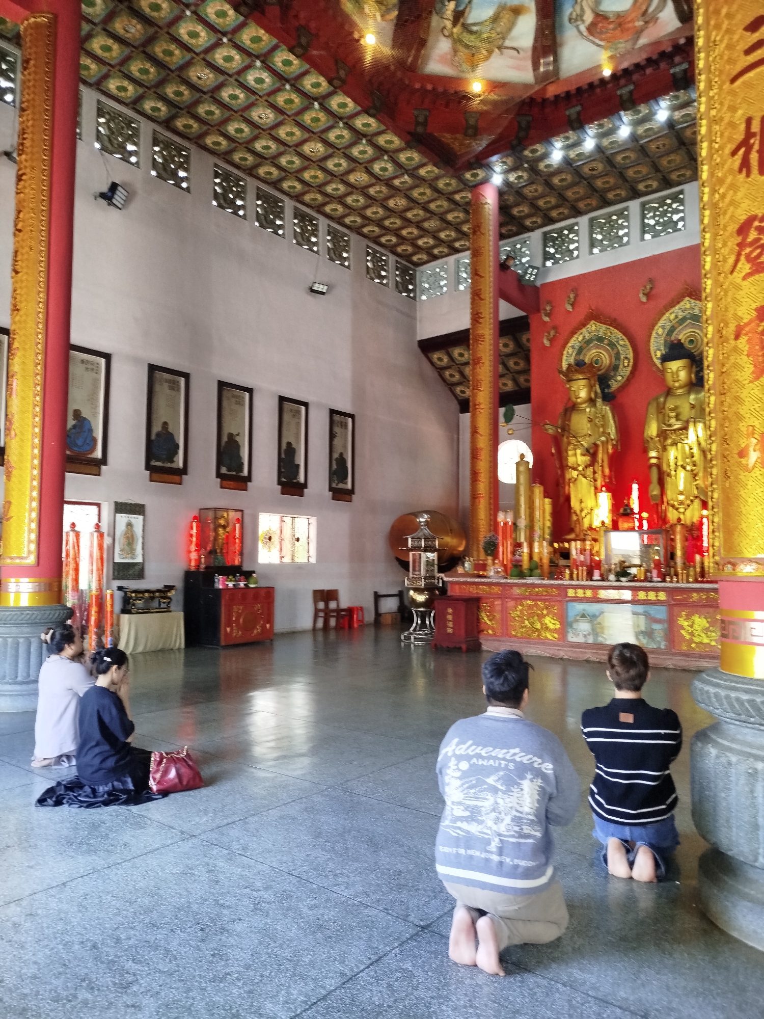 The image shows people worshiping in a temple in Da Lat, Vietnam. Inside a large hall, several people kneel on the grey stone floor, facing an altar adorned with golden statues and various offerings. The altar sits against a red backdrop, with ornate gold detailing on the surrounding pillars. The ceiling is elaborately decorated with a patterned design, and framed portraits line the walls. Lighting fixtures are visible near the ceiling, and the overall environment suggests a serene, reverential atmosphere.