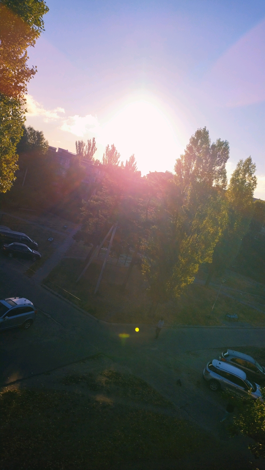 In Horishni Plavni, Ukraine, a scene unfolds with the sun setting or rising, casting a strong, pinkish-orange glow across the sky and through the leaves of trees. The light creates a lens flare effect. Below, apartment buildings are visible, partially obscured by the trees. Several cars are parked on a paved area that appears to be a street or parking lot, including a silver station wagon in the foreground and a dark-colored sedan further back. A person is walking across the paved area, illuminated by a yellow circular light source on the ground. The environment appears to be a residential area, with a mixture of buildings, trees, and paved surfaces.