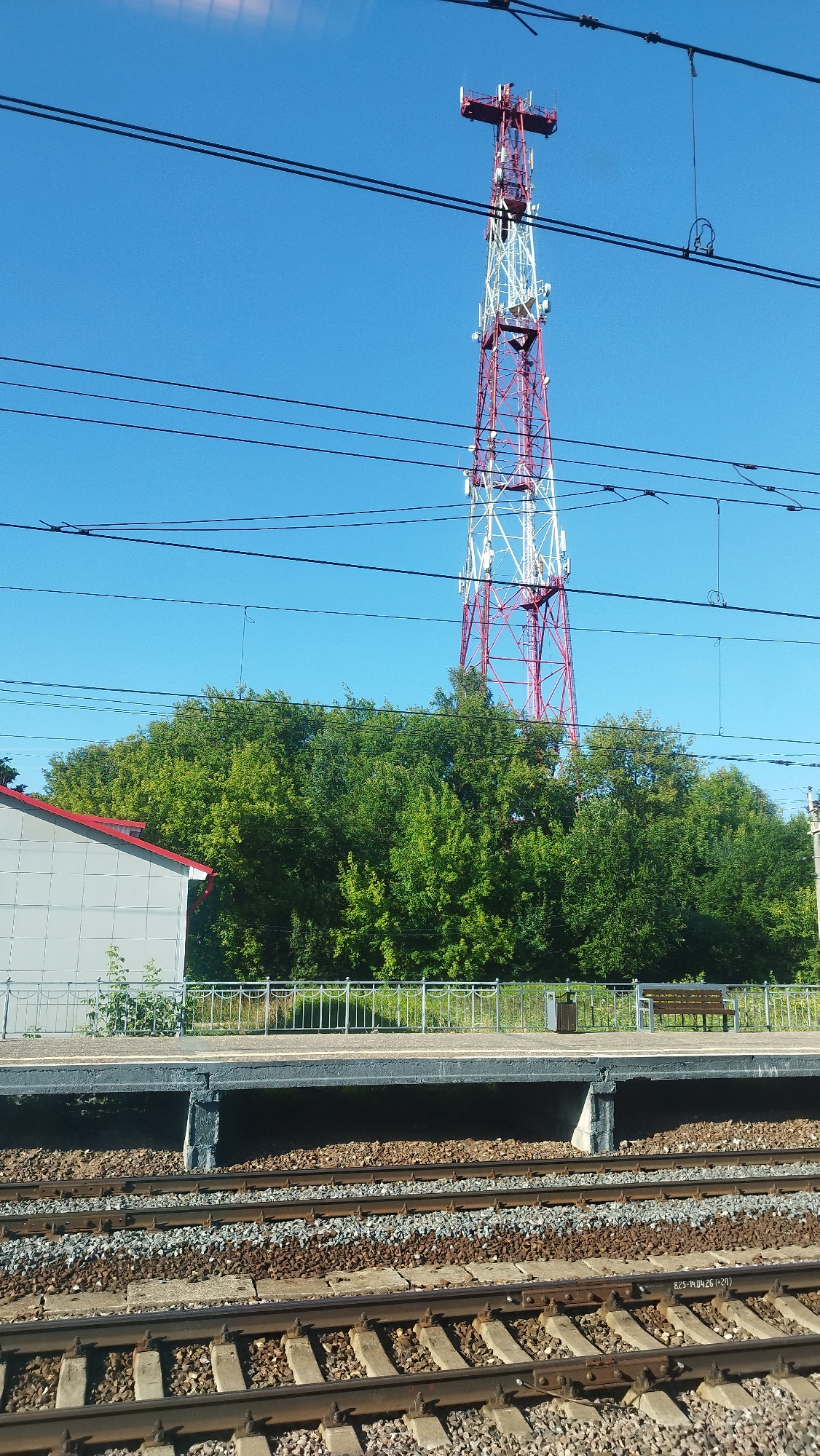 A low-angle, full-frame outdoor shot depicts a railway platform and tracks against a clear blue sky. Dominating the upper half of the frame is a tall, red and white lattice telecommunications tower. Several parallel black cables crisscross the sky in front of the tower. Below the cables and tower, a dense line of lush green trees forms a backdrop. In the foreground, a gray building with a red-tiled roof is partially visible on the left. A metal railing with a light-colored platform extends horizontally across the middle ground. On the platform, a dark-colored trash receptacle and a wooden bench are discernible. The bottom portion of the image shows a detailed view of multiple sets of railway tracks, characterized by ballast, sleepers, and rails.