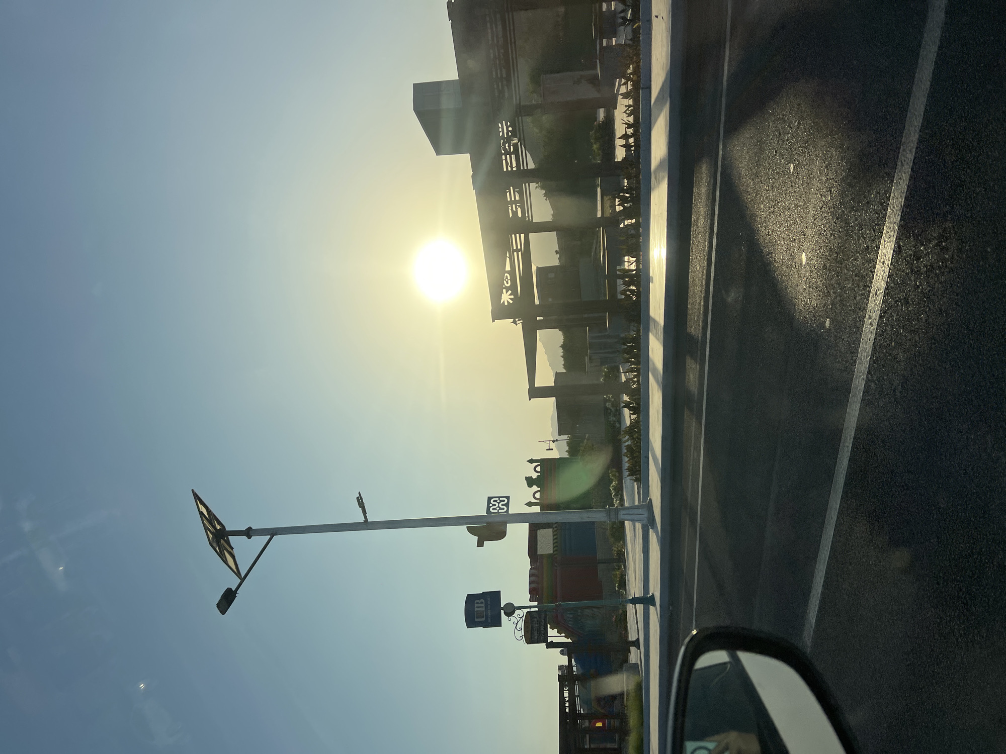 The image shows a sun-drenched street scene in Sharm El Sheikh, Egypt. The sun is low in the sky, casting a warm, golden glow and creating long shadows. In the foreground, a car's side mirror is visible on the left, reflecting the driver's hand holding a phone. The street itself is asphalt with white lane markings. A sidewalk lines the street, featuring a solar-powered street lamp with a rectangular solar panel and a light fixture. On the sidewalk, there are several signs: one with the logo "UB" and text "THE WAY OF WORT", another with Arabic script, and a third with a stylized symbol that appears to be "52". Beyond the sidewalk, there is a landscaped area with low green plants and a pergola structure with decorative cutouts. In the background, a modern building with a dark facade stands against the clear blue sky. The overall impression is one of a modern urban development bathed in the light of a setting or rising sun.