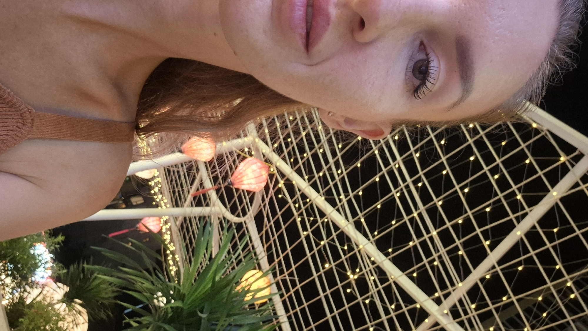 The image captures a close-up, partial view of a fair-skinned woman with reddish-brown hair, her face turned slightly to the left. Her left eye, framed by dark mascara, is visible, along with her nose and lips, which appear to have a light pink gloss. She is wearing a tan, ribbed camisole.
The background suggests an outdoor setting at night, likely in Đà Nẵng, Vietnam, given the presence of decorative elements often seen in this city. A white lattice structure, illuminated by numerous small, warm-toned lights, forms an overhead canopy. Dangling from this structure are several glowing red lanterns, some appearing spherical and others more elongated, typical of Asian festival decorations. In the lower right portion of the frame, a lush green plant with long, frond-like leaves is partially visible, adding a touch of nature to the scene. The overall ambiance suggests a festive or decorative outdoor space, possibly a restaurant, market, or entertainment area, illuminated by artificial lights and lanterns.