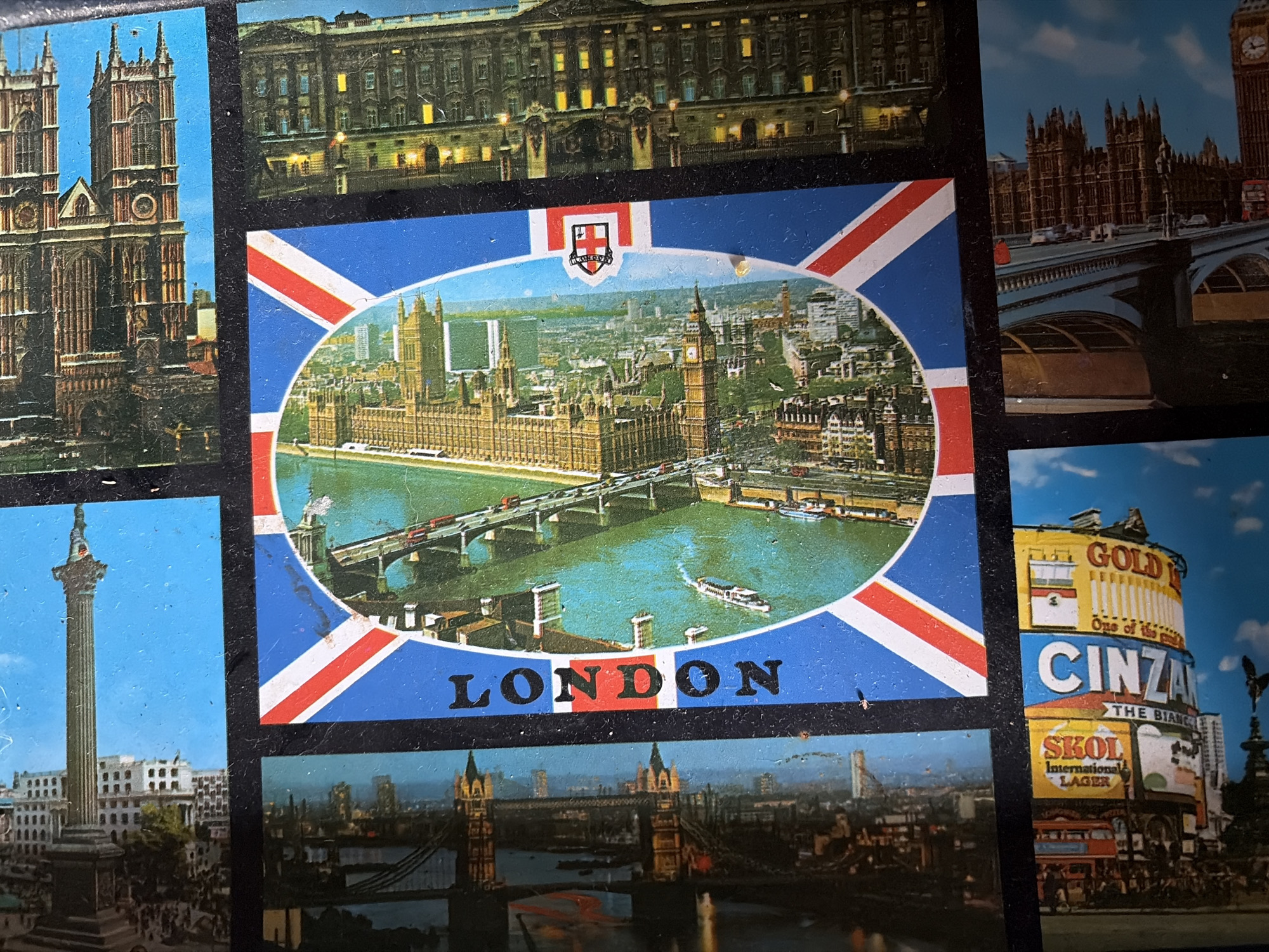 The image displays a collection of London postcards, some featuring iconic landmarks like the Houses of Parliament and Nelson's Column. One postcard has "LONDON" written vertically. Another shows a view of the Houses of Parliament from across the River Thames, with a bridge in the foreground. There are also images of Big Ben and other architectural structures. The bottom section of the image shows a postcard with advertisements for "SKOL International Lager," "CINZANO," and "GOLD." This image is not of Malyshevo, Russia.
